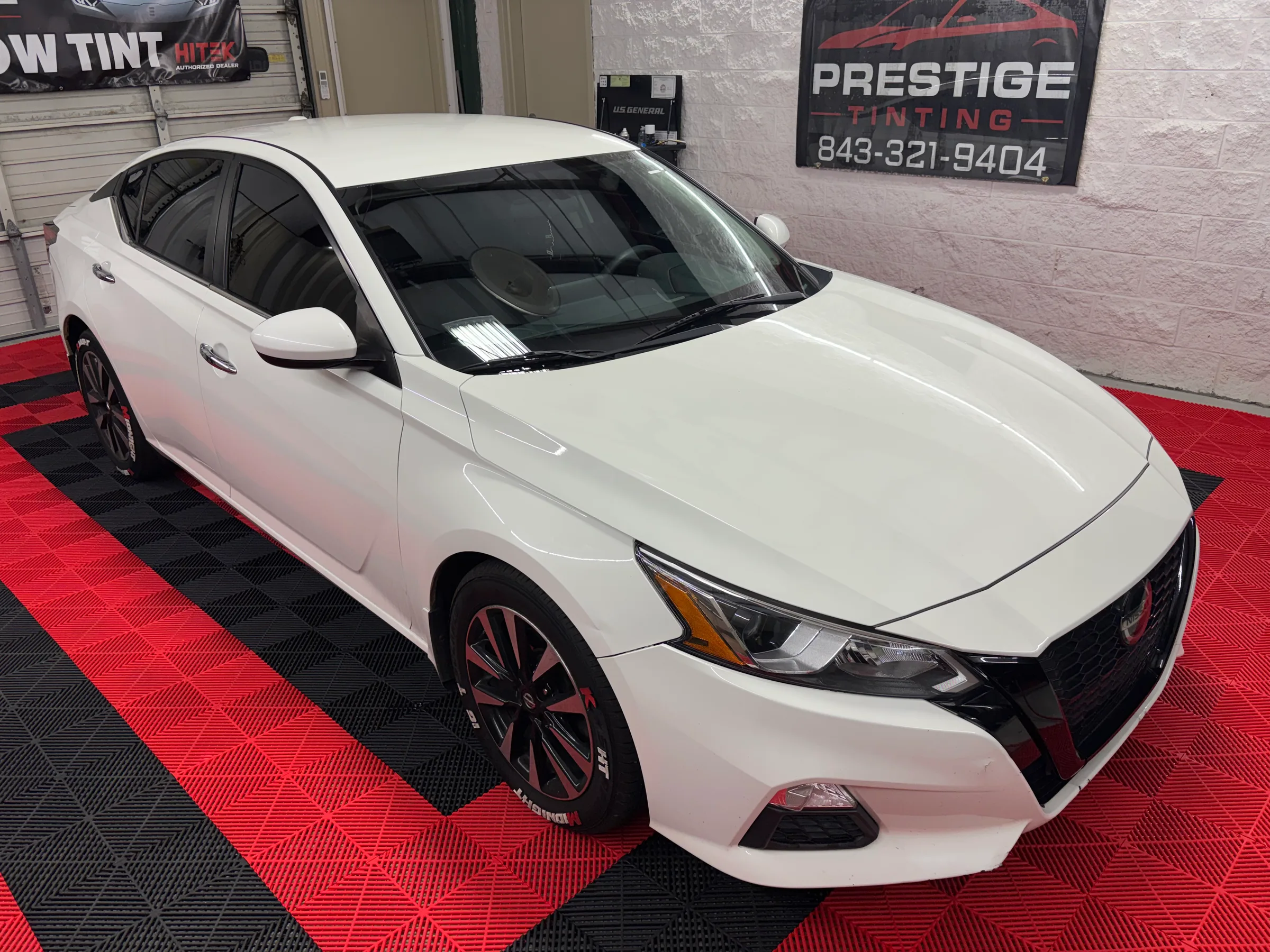 A white Nissan Altima with dark window tint displayed in a shop with red and black floor mat, Prestige Tinting signage visible behind the vehicle.