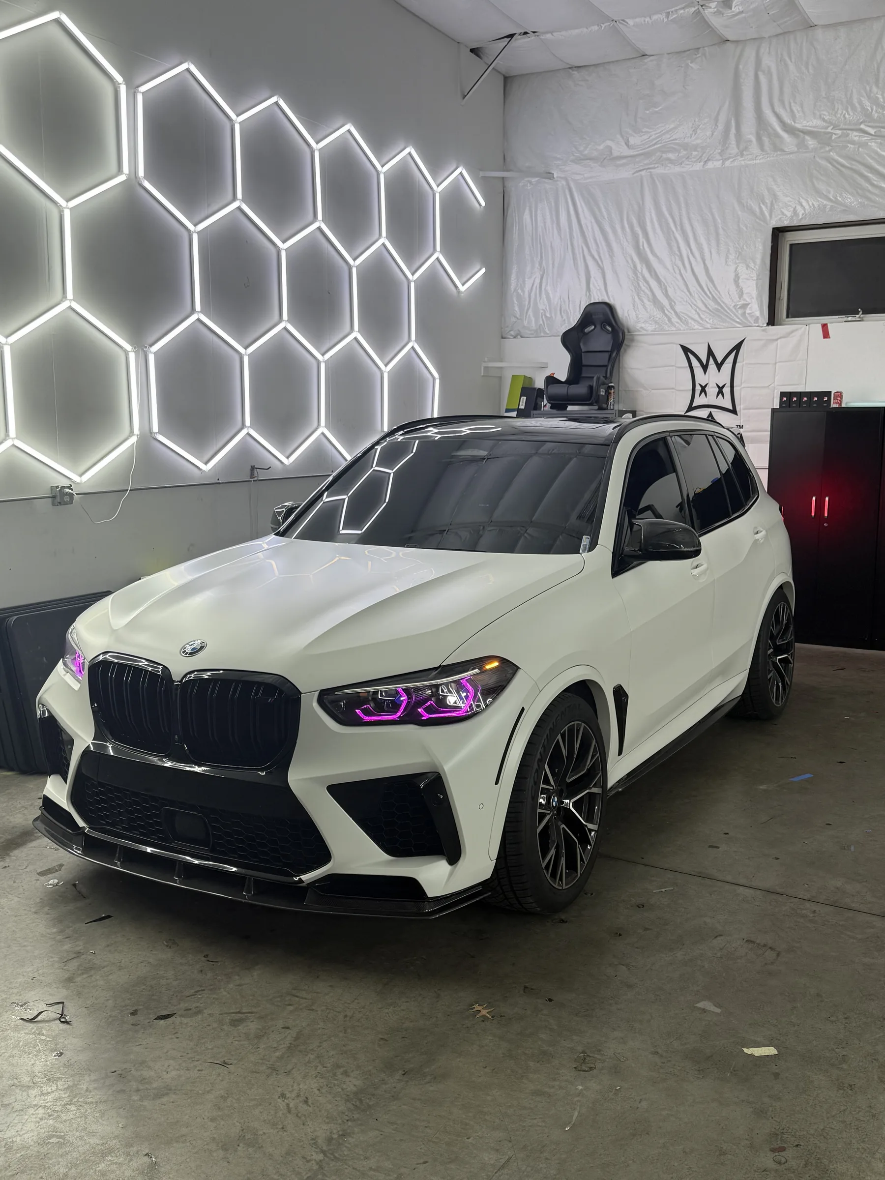 White BMW X5 with purple-tinted headlights in a shop bay with neon hexagon wall decor.