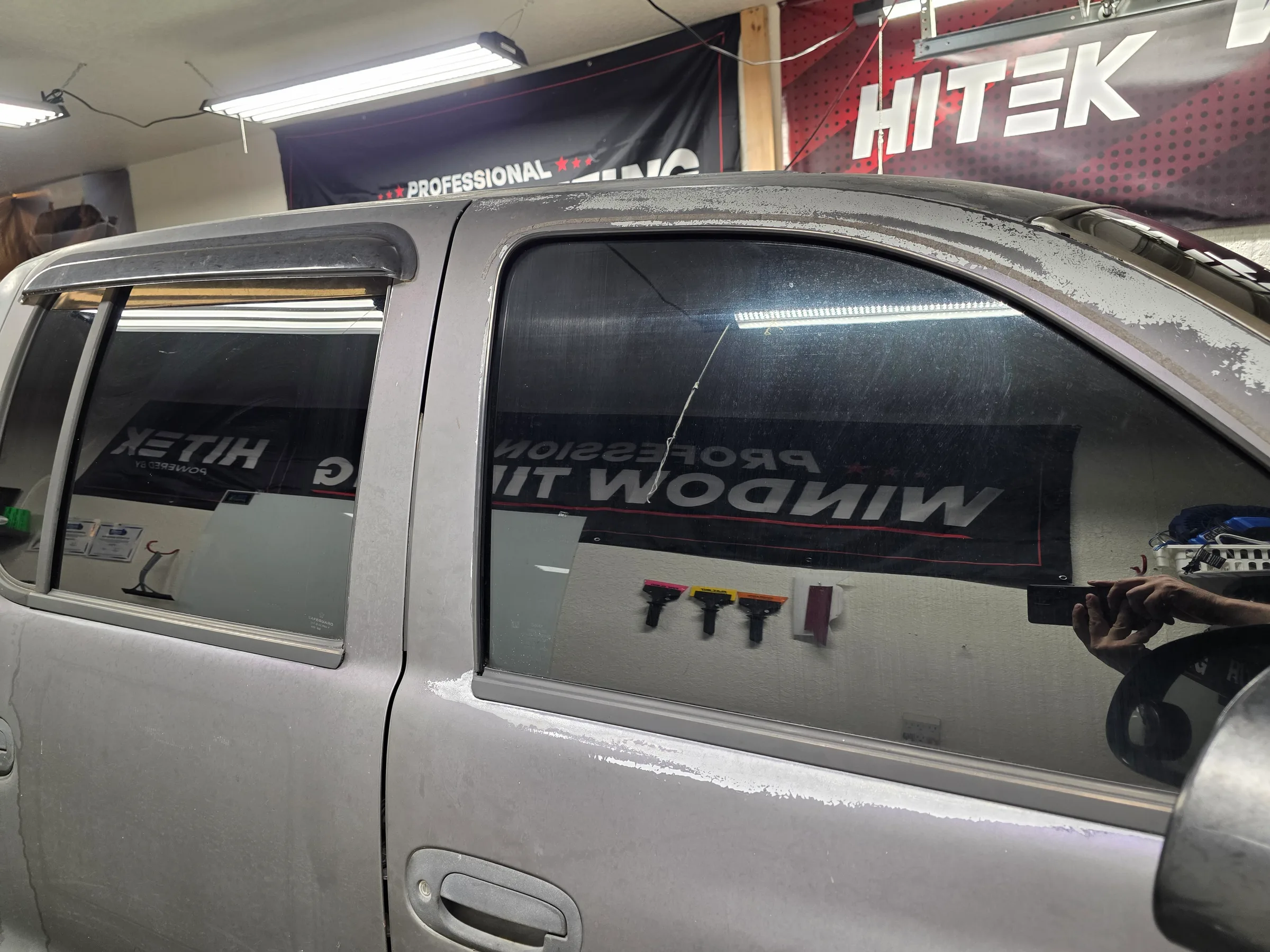 A white pickup truck with dark window tint being applied to the rear windows inside a professional tinting shop.