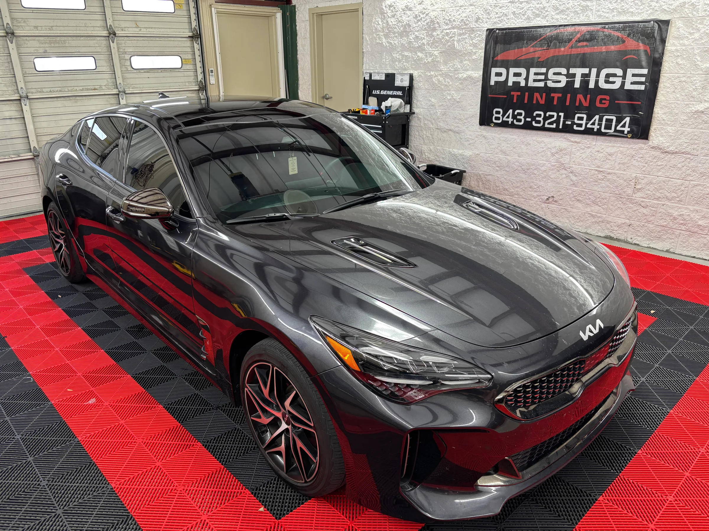 A dark gray Kia Stinger sedan with tinted windows parked in a professional workshop bay with red and black flooring and a Prestige Tinting sign displayed on the wall.