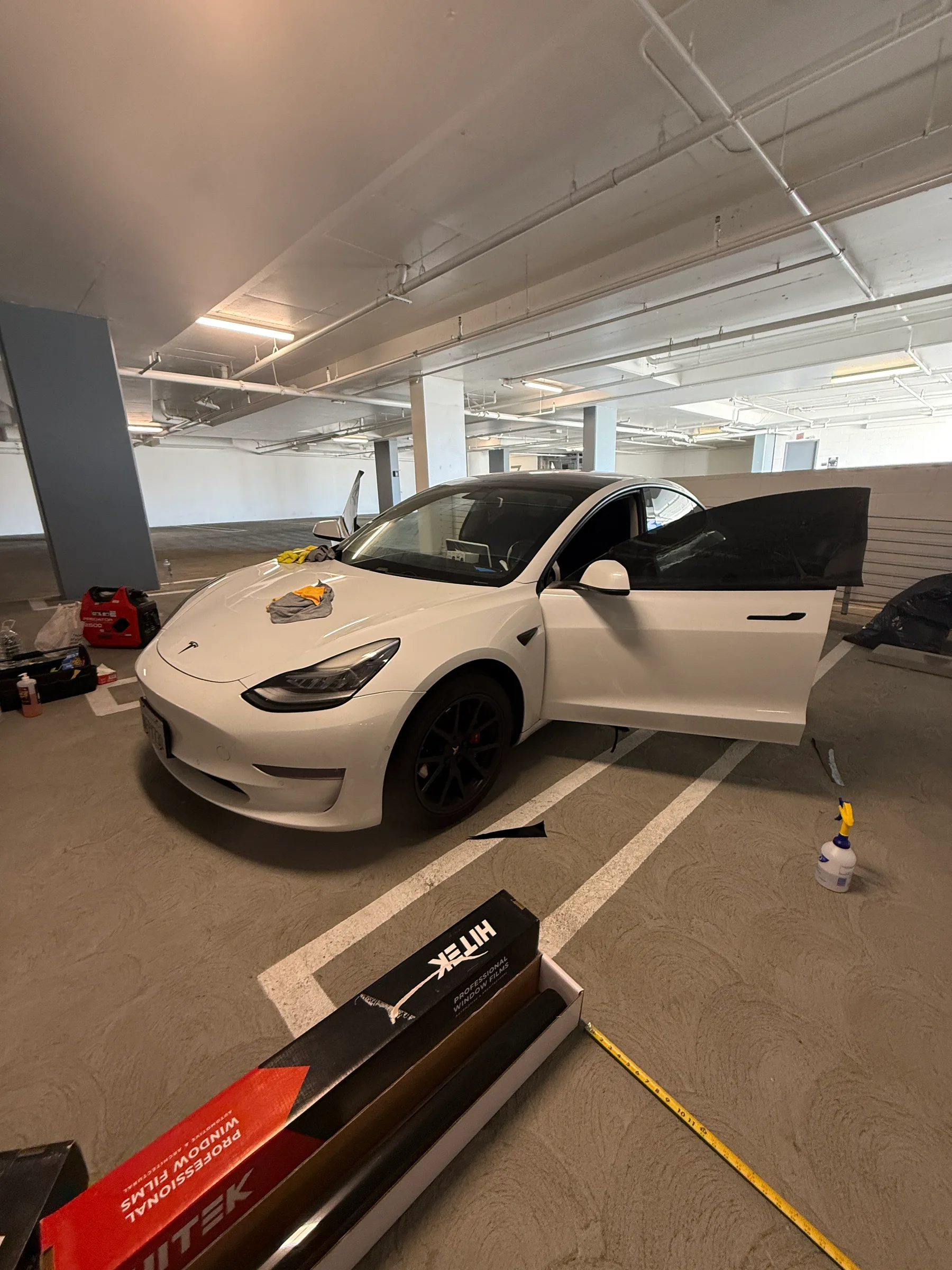 White Tesla Model 3 with door open in a tint shop bay, surrounded by window tint rolls and application tools.