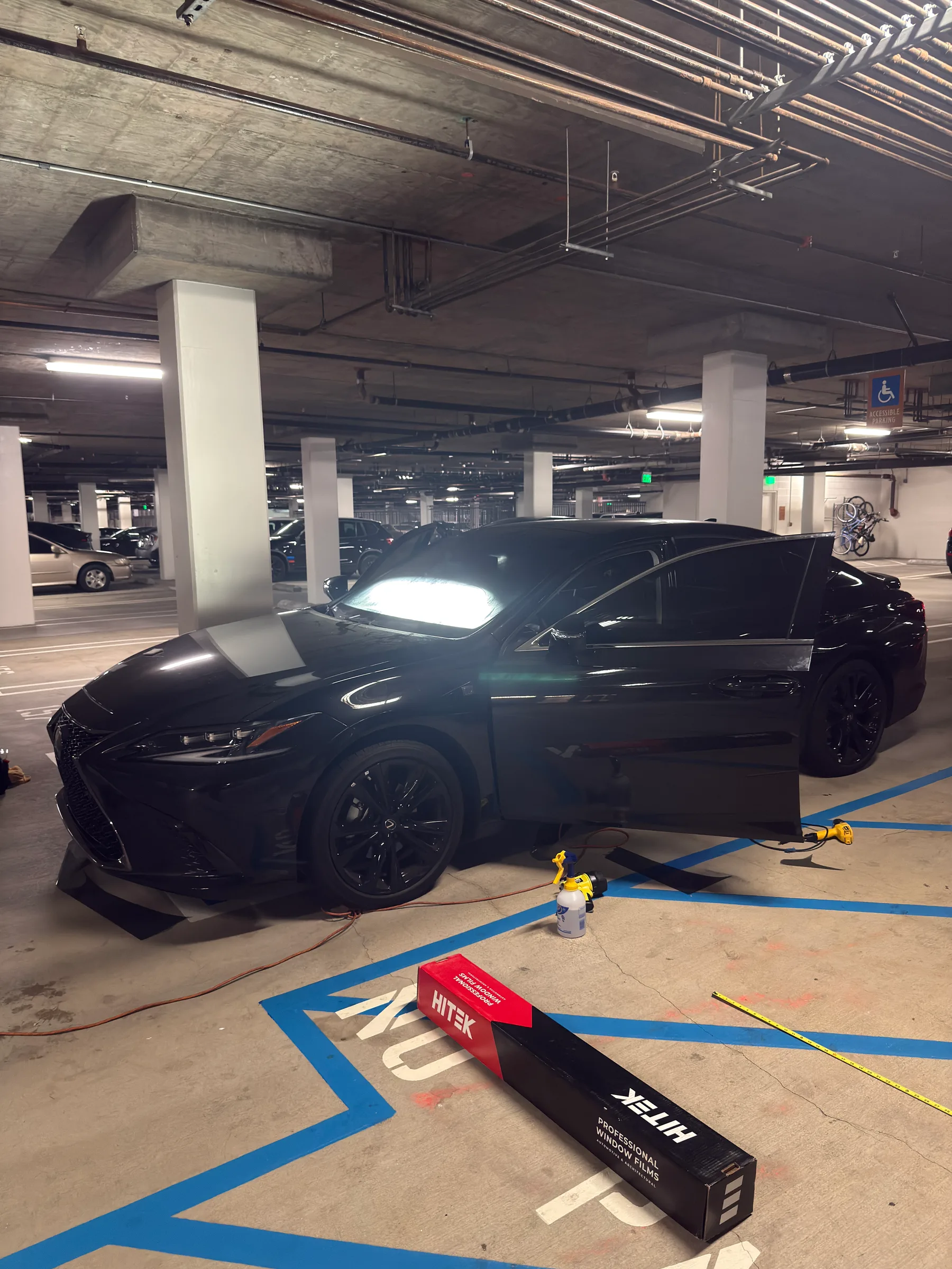 A black Lexus sedan with an open driver door receives window tinting in an indoor parking garage, with tinting materials and equipment visible on the ground.