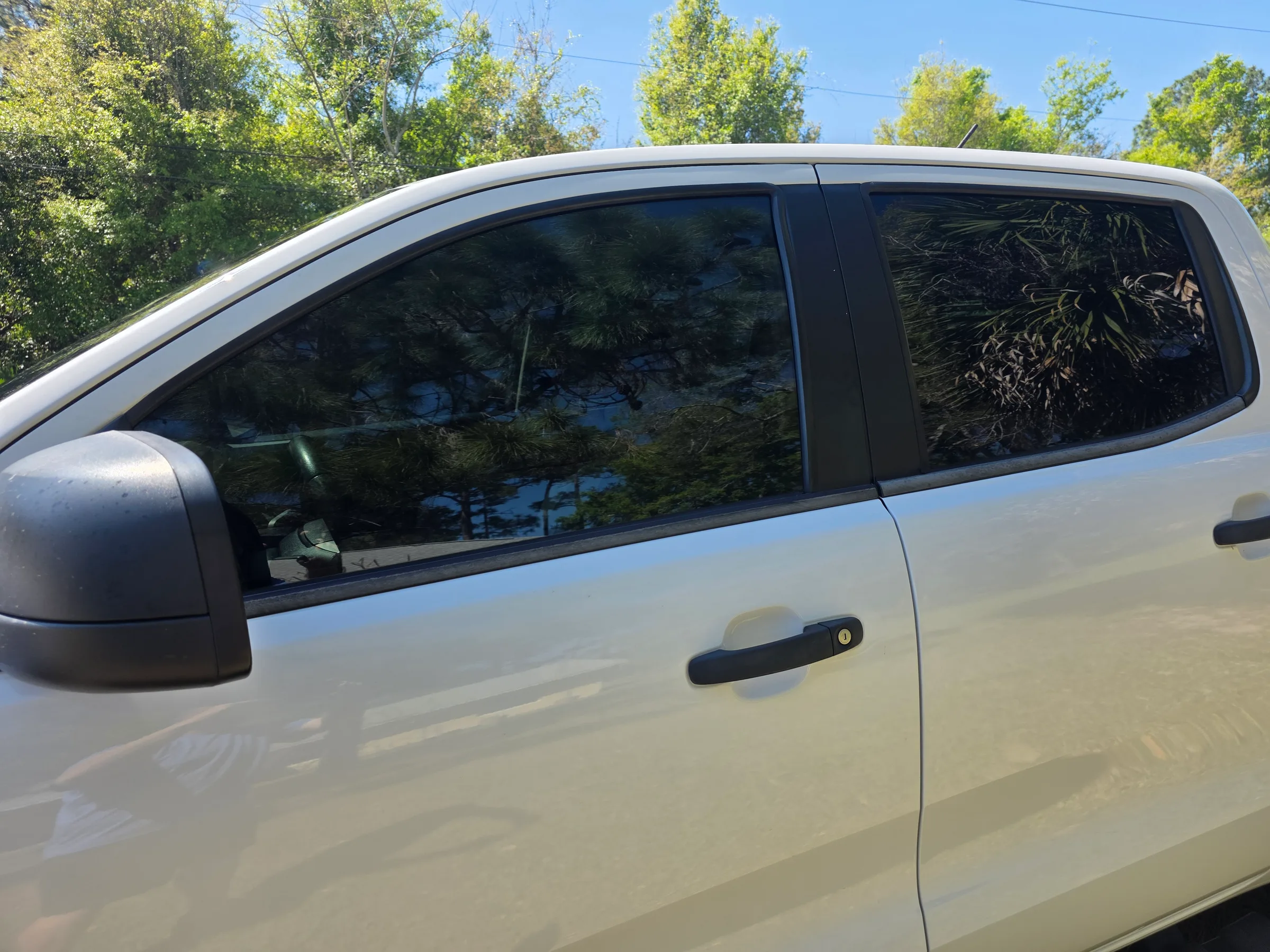 Side view of a white pickup truck with dark window tint applied to the rear cab windows, parked outdoors with trees in the background.