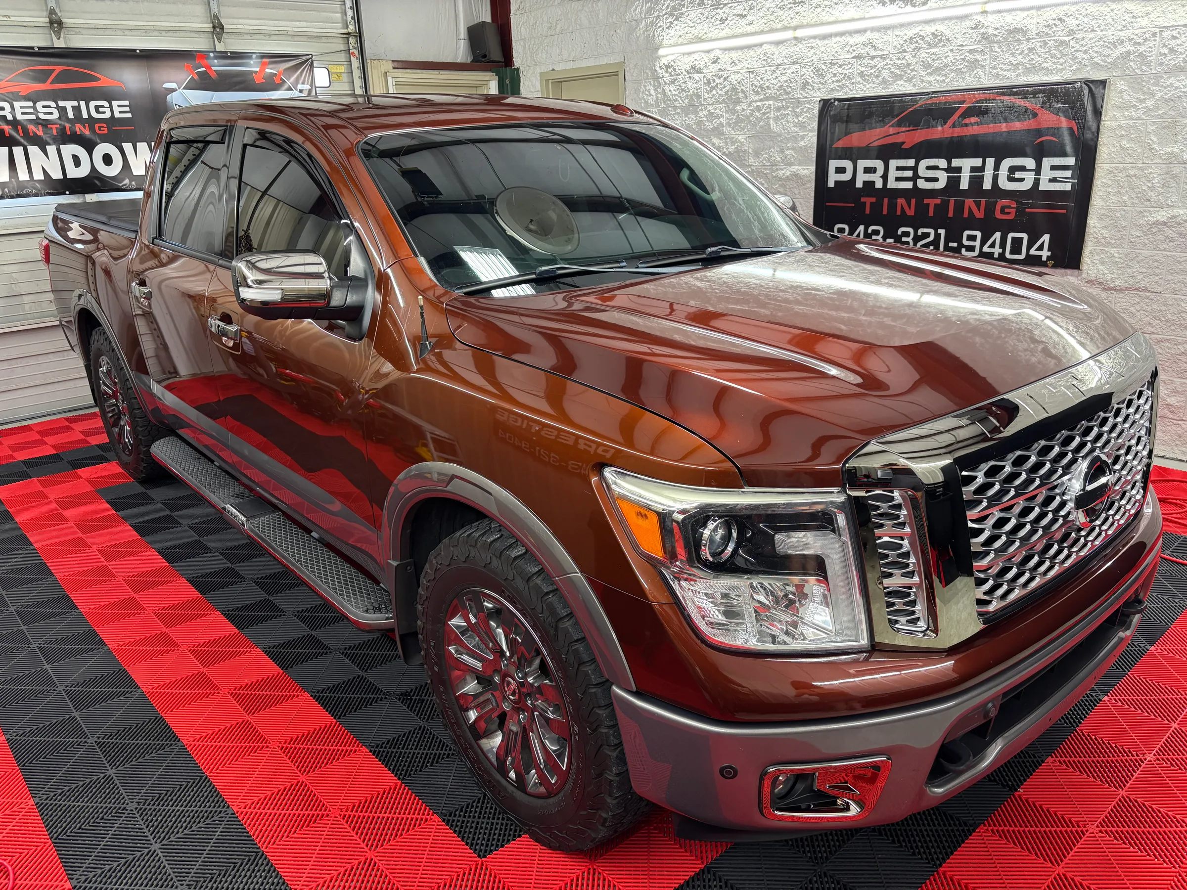 A burgundy Nissan Titan pickup truck with window tint applied, displayed in a showroom with red and black flooring and Prestige Tinting signage.