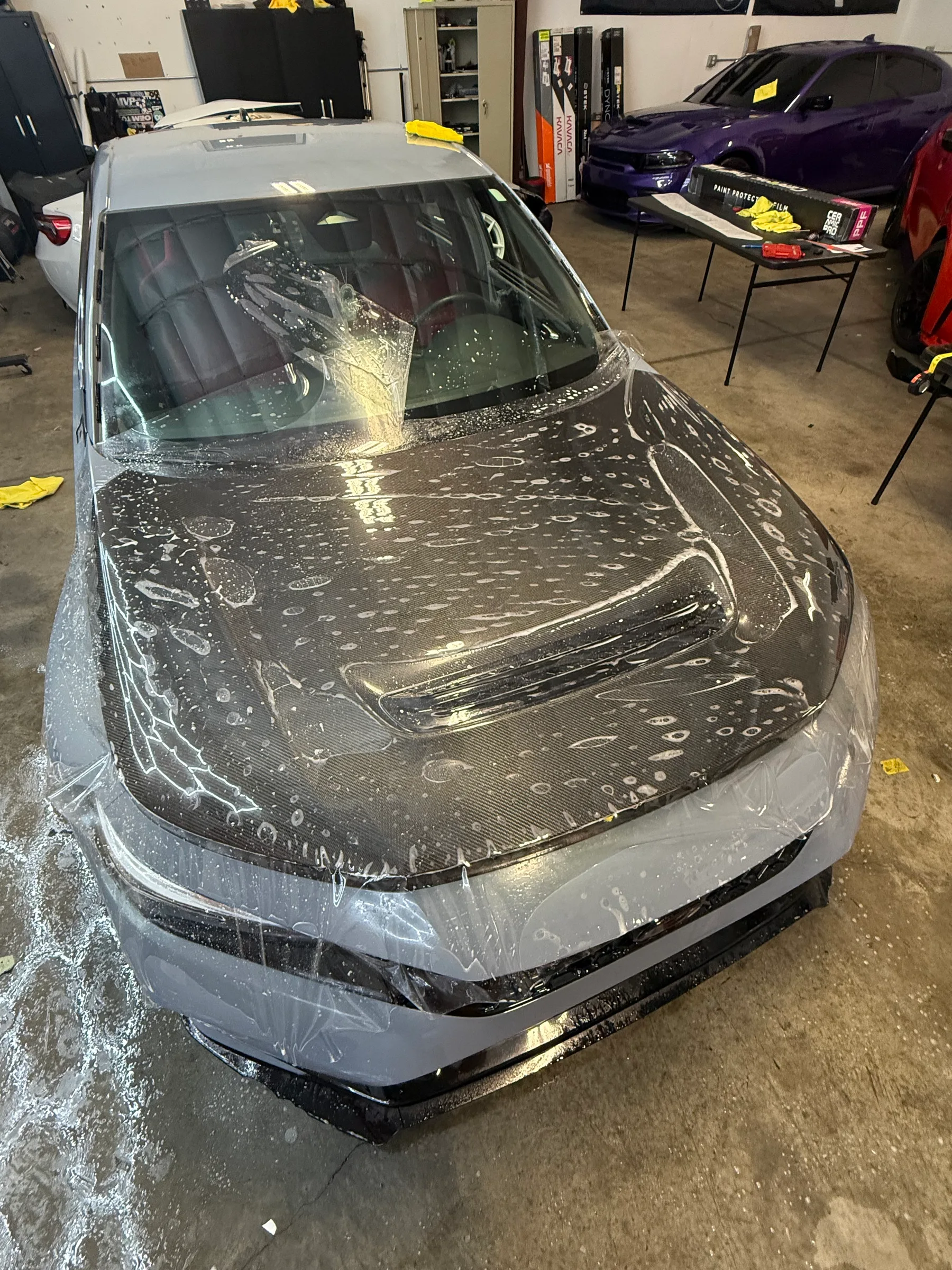 A silver sedan receiving paint protection film application, shown in a shop with the hood and front end covered in clear protective material.