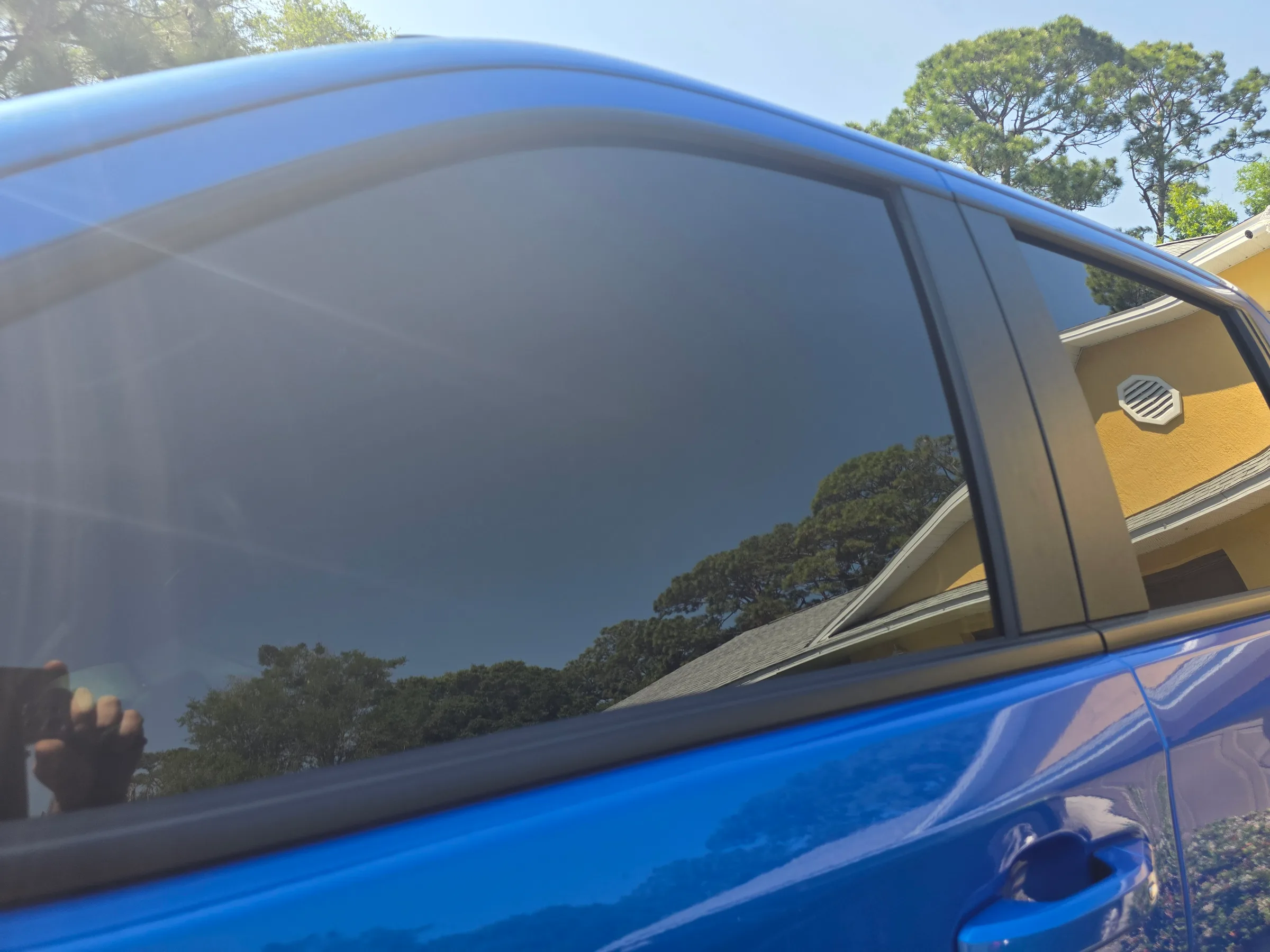 Close-up view of a blue vehicle's side window with dark tinted film applied, showing the tinted glass against a sunny outdoor setting.