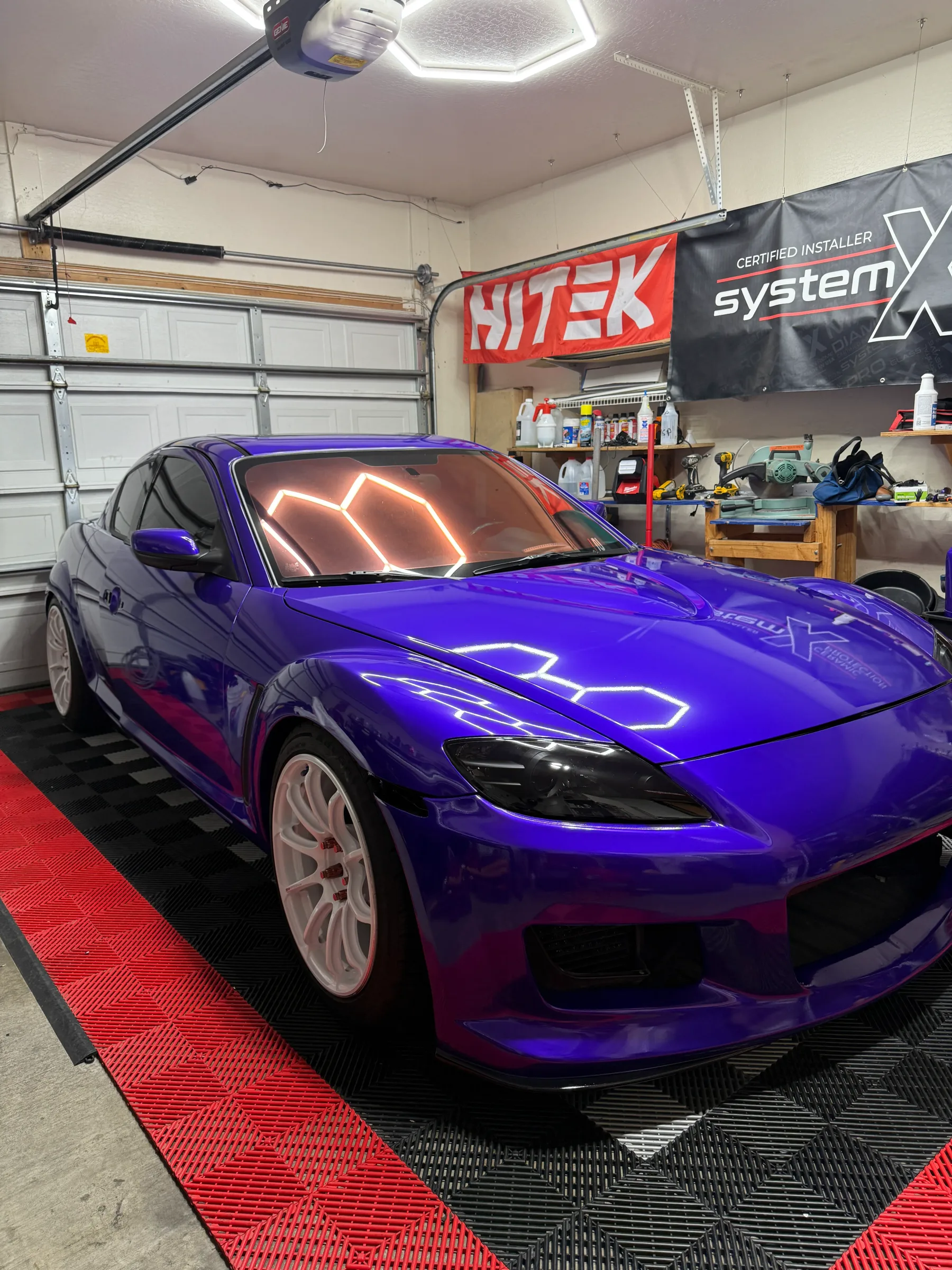 A vibrant purple Mazda RX-8 with neon headlight and side marker accents parked in a professional installation bay, displaying window tint and vehicle customization work.