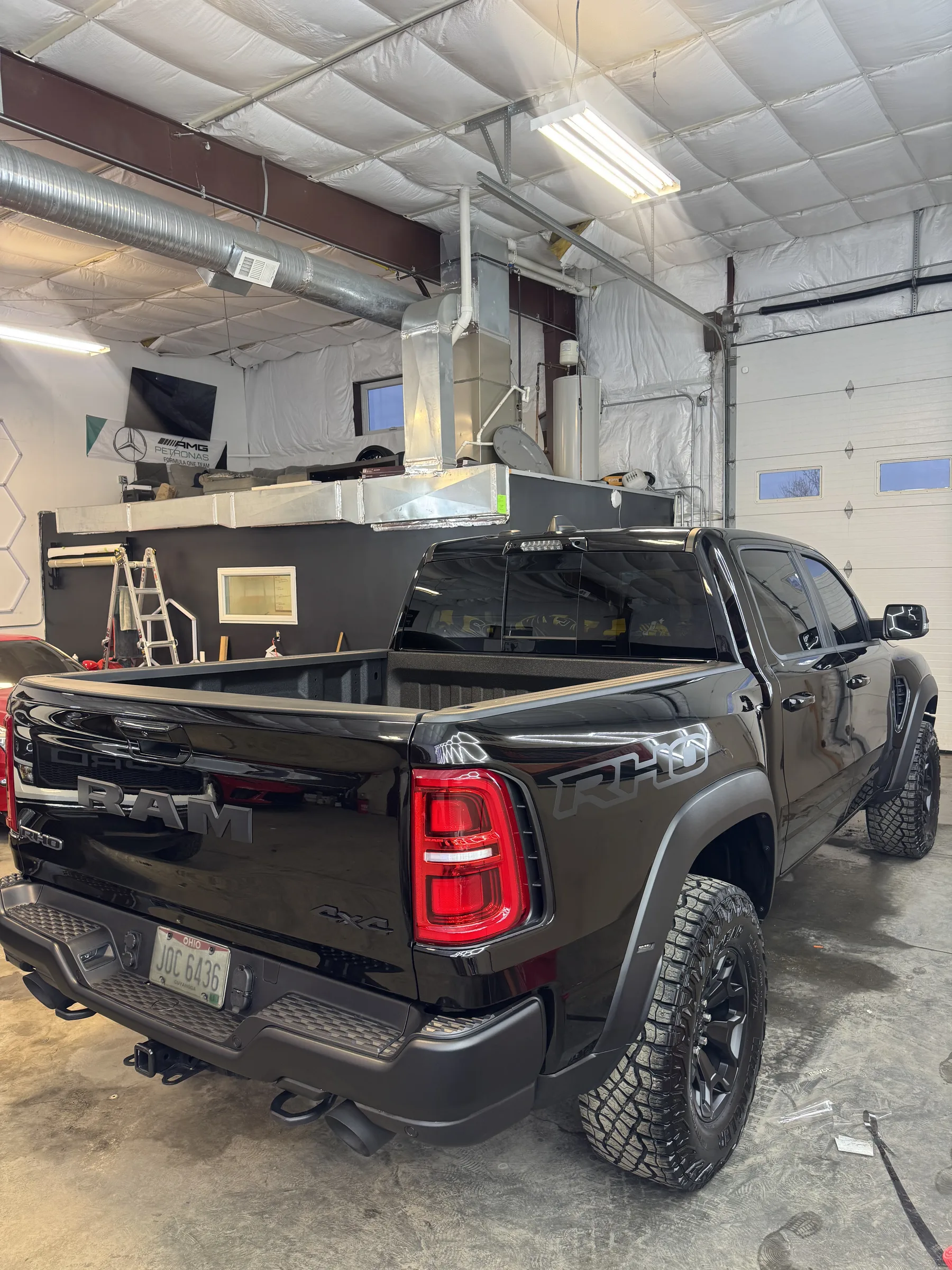 Black RAM pickup truck in a professional shop receiving paint protection film installation, shown from the rear three-quarter angle with garage bay doors and work station visible.