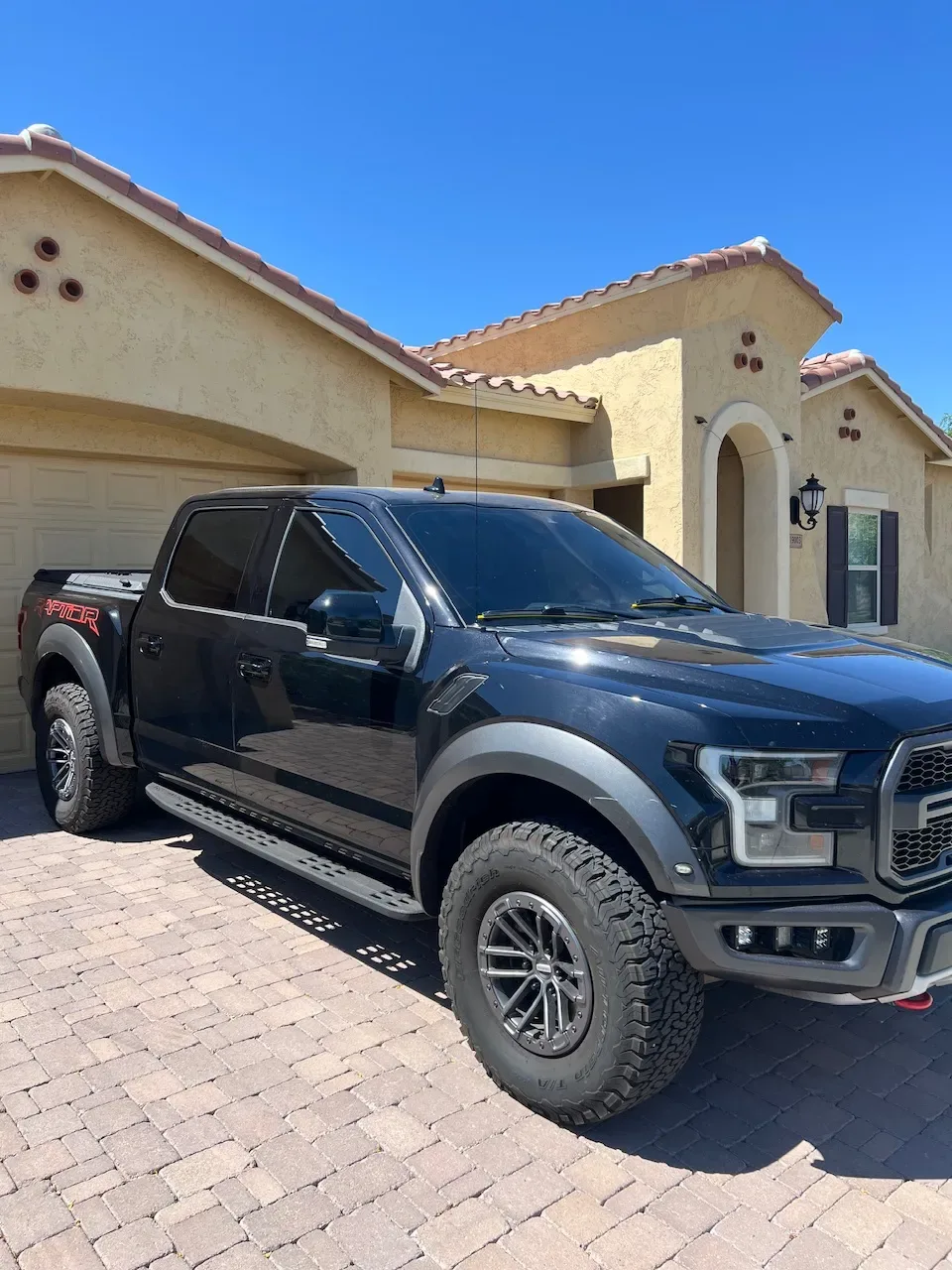 A black Ford Ranger pickup truck with dark window tint parked in the driveway of a tan stucco home under clear Arizona skies.