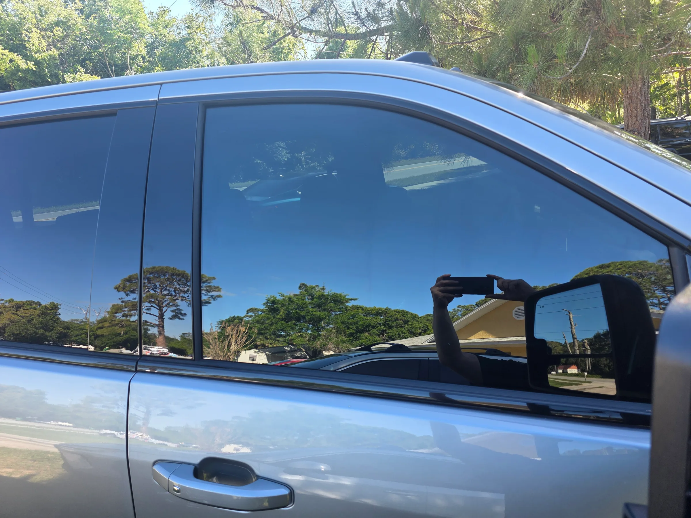 A blue van with dark window tint applied to the side and rear windows, parked outdoors with trees visible in the reflection.