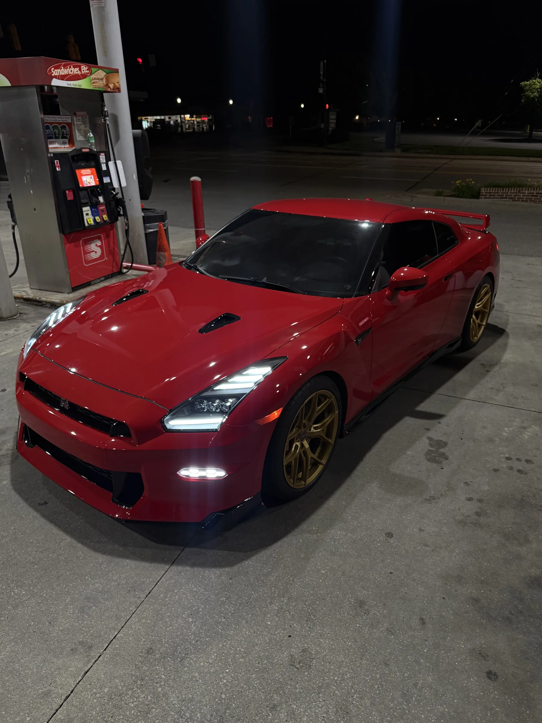 A bright red Nissan GT-R with gold wheels parked at a gas station at night, showcasing its glossy finish and tinted windows.