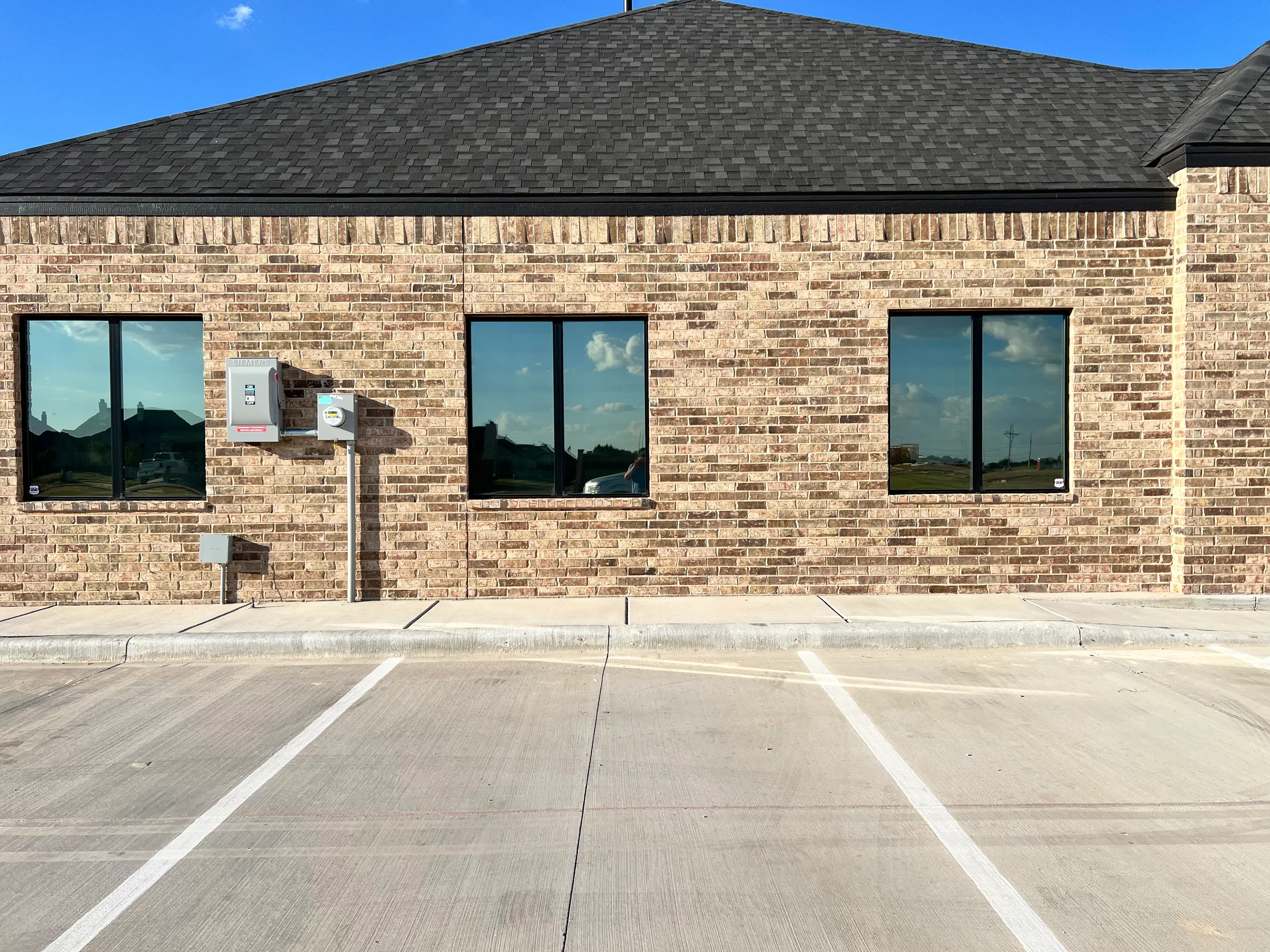 A brick commercial building with three large windows tinted with dark film, reflecting the sky and surrounding landscape.