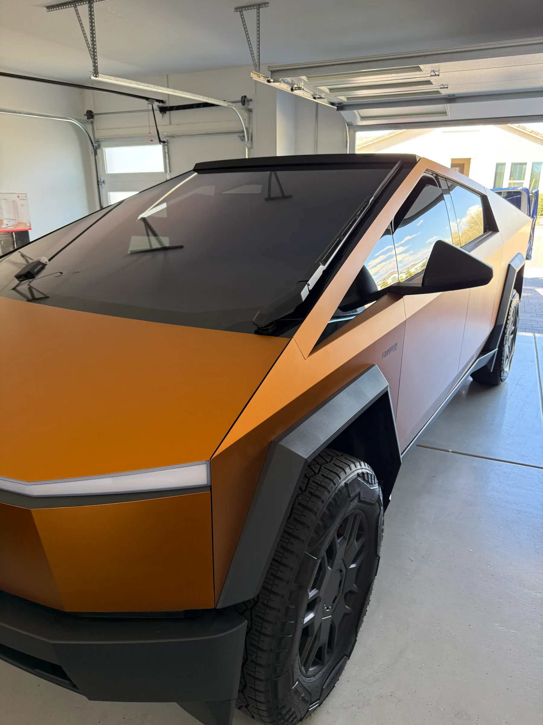An orange and black Tesla Cybertruck with dark window tint applied to the side windows, parked in a professional auto shop with garage doors.