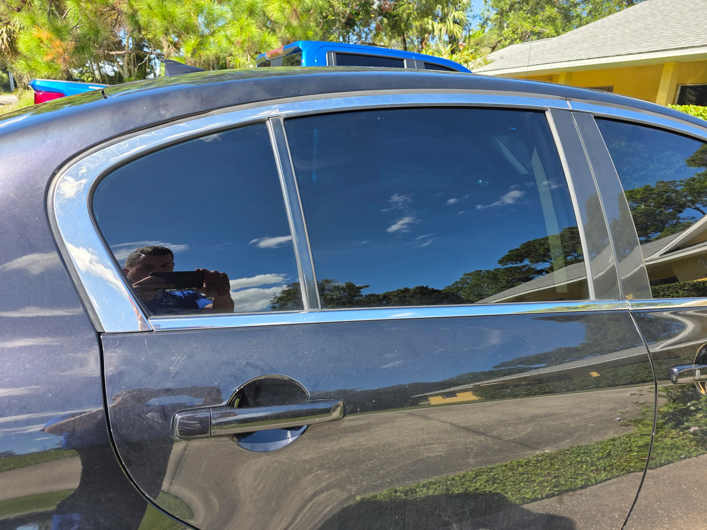 A blue SUV with dark window tint applied to the side windows, parked in a residential driveway with trees and a yellow house visible in the background.