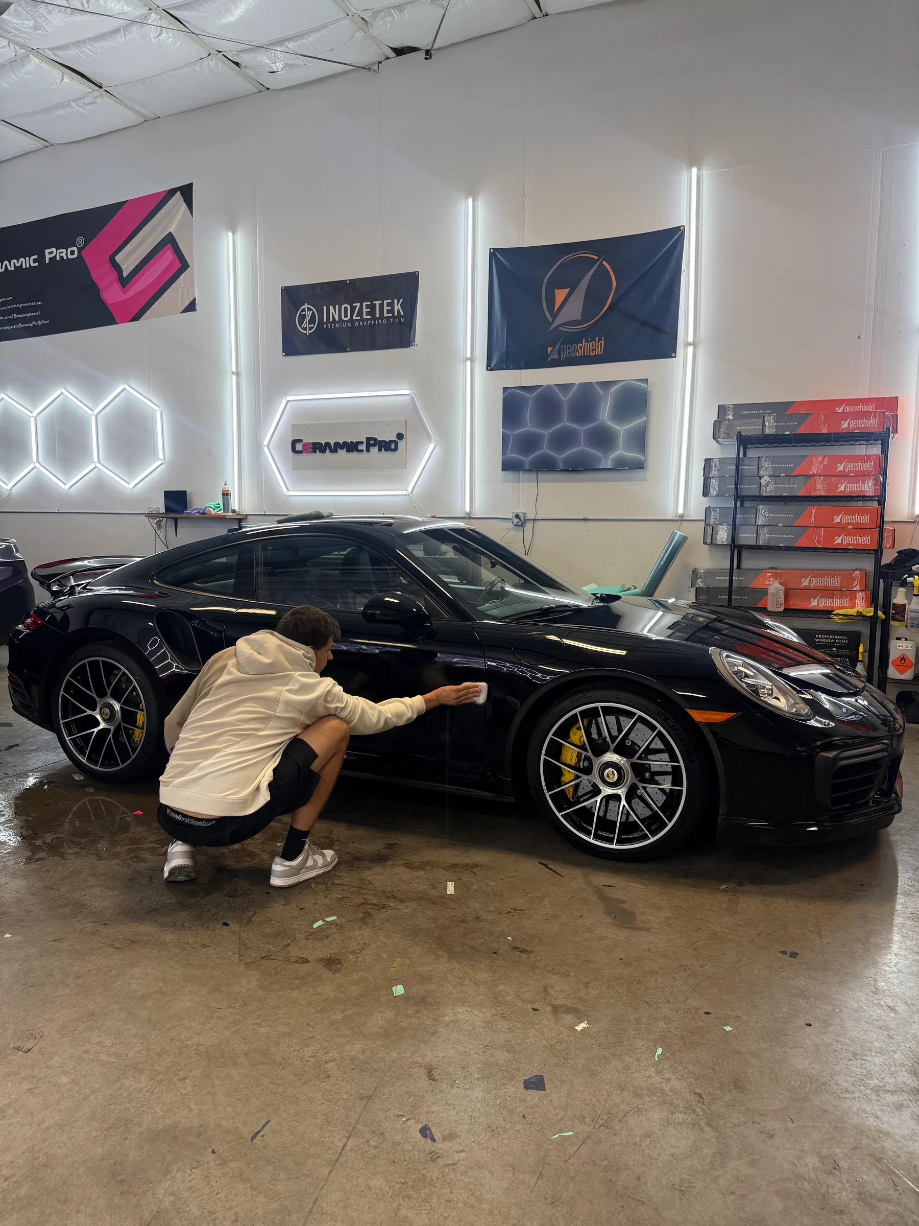 A technician applies protective film to the side of a black Porsche sports car in a shop equipped with brand logos and neon signage.