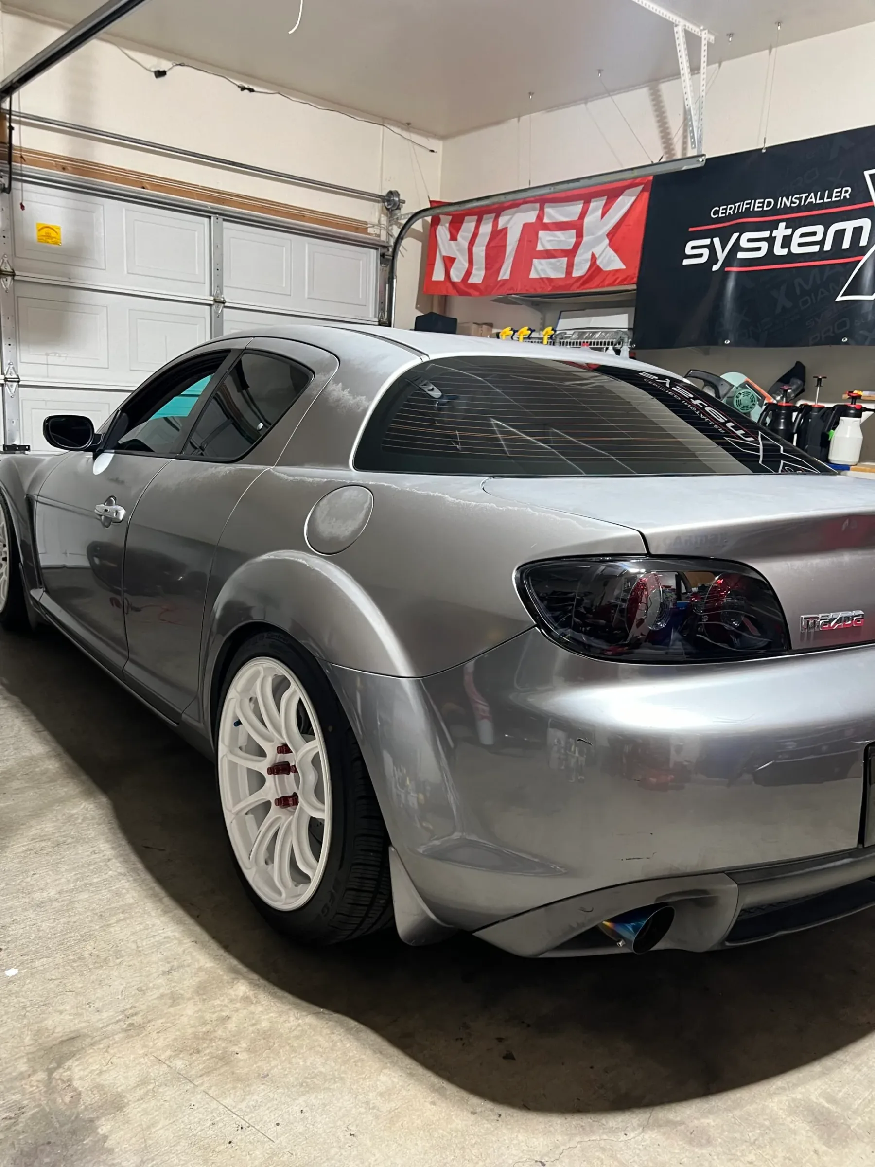 Silver Mazda RX-8 with dark window tint installed, parked in a professional shop with branding visible in the background.