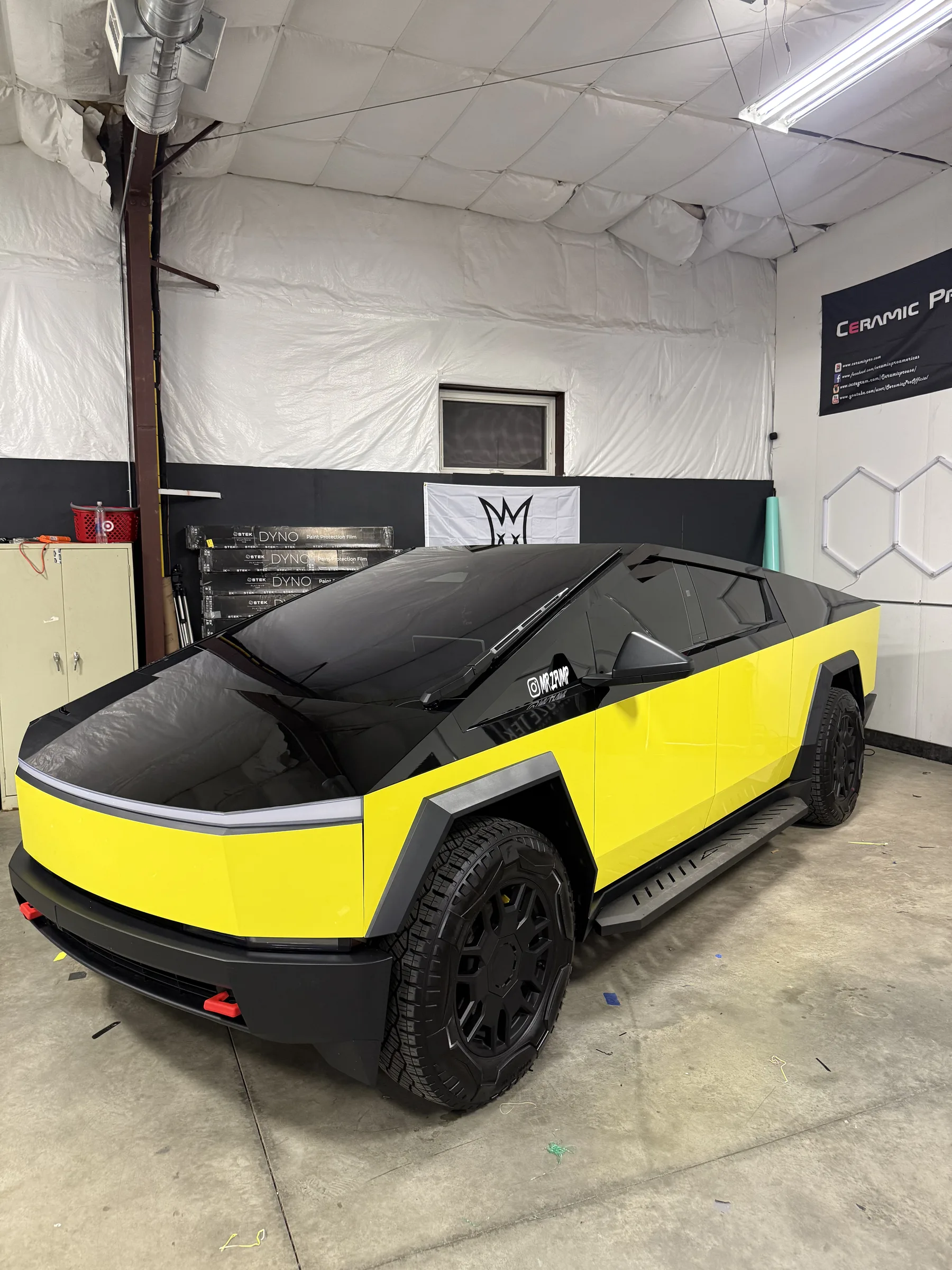 A Tesla Cybertruck with a two-tone yellow and black vinyl wrap parked in a professional auto shop in Berea, OH.