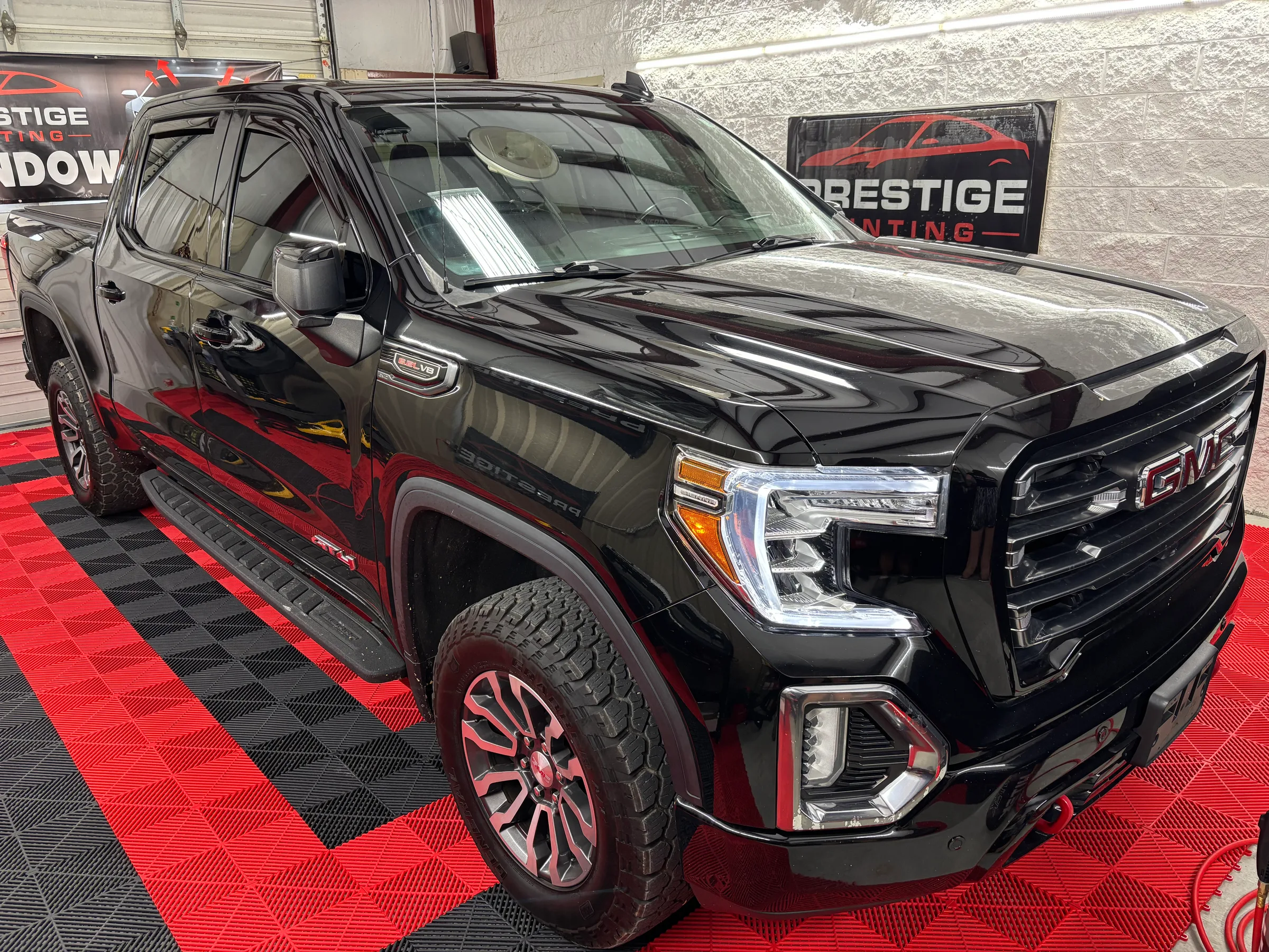 A black GMC Sierra pickup truck with tinted windows displayed in a shop showroom, featuring custom red and black wheels on a red and black checkered floor.
