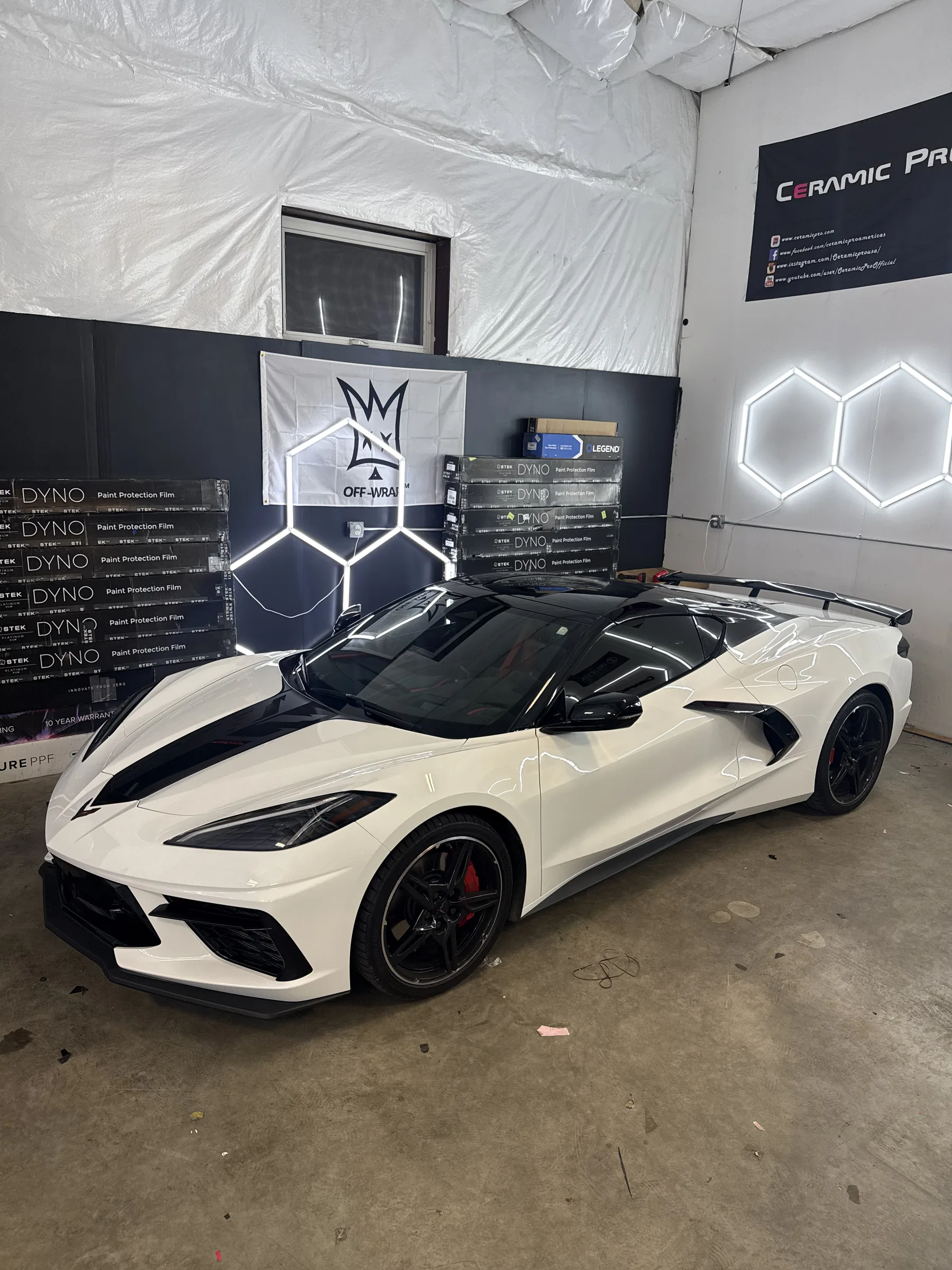 A white Corvette sports car with black racing stripes displayed in a shop bay surrounded by paint protection film product boxes and branding signage.