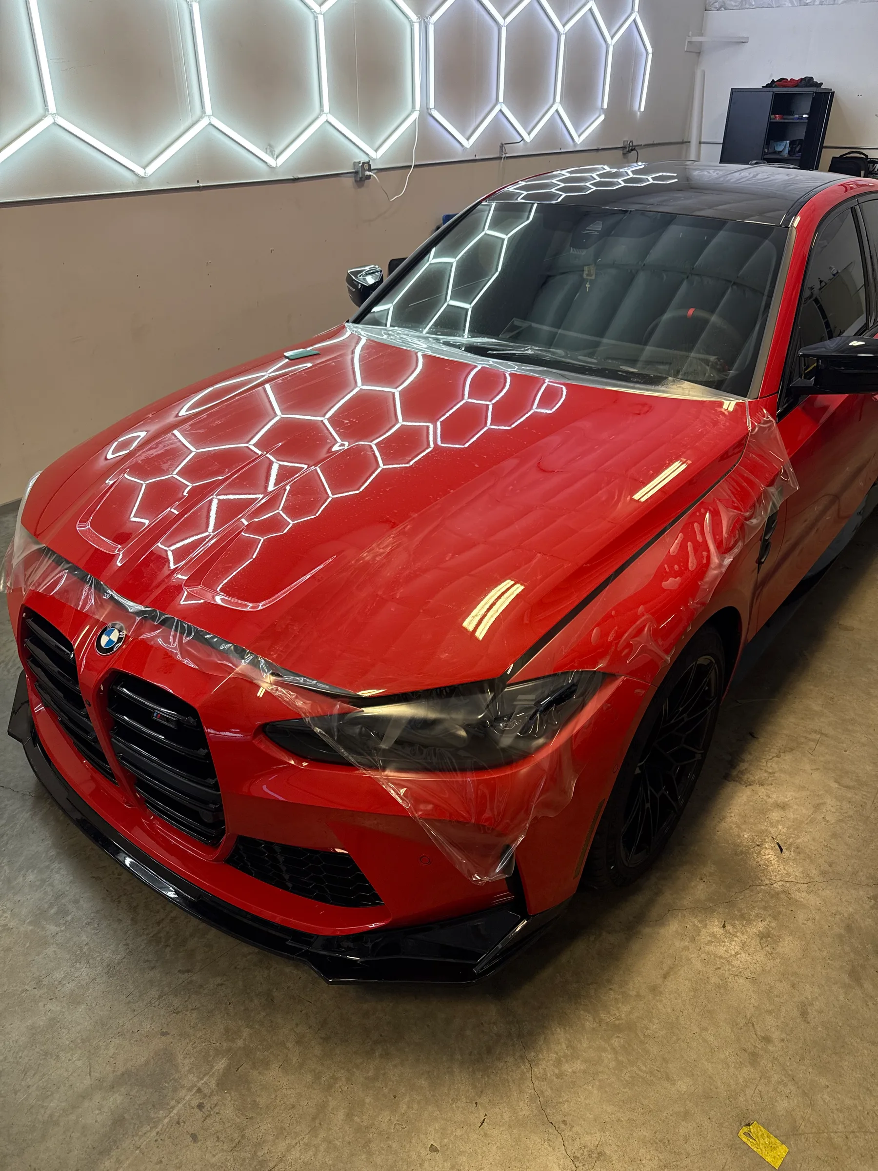 A red BMW sedan with protective film being applied to the hood and windshield, showing the transparent hexagonal pattern of paint protection film in a shop interior.