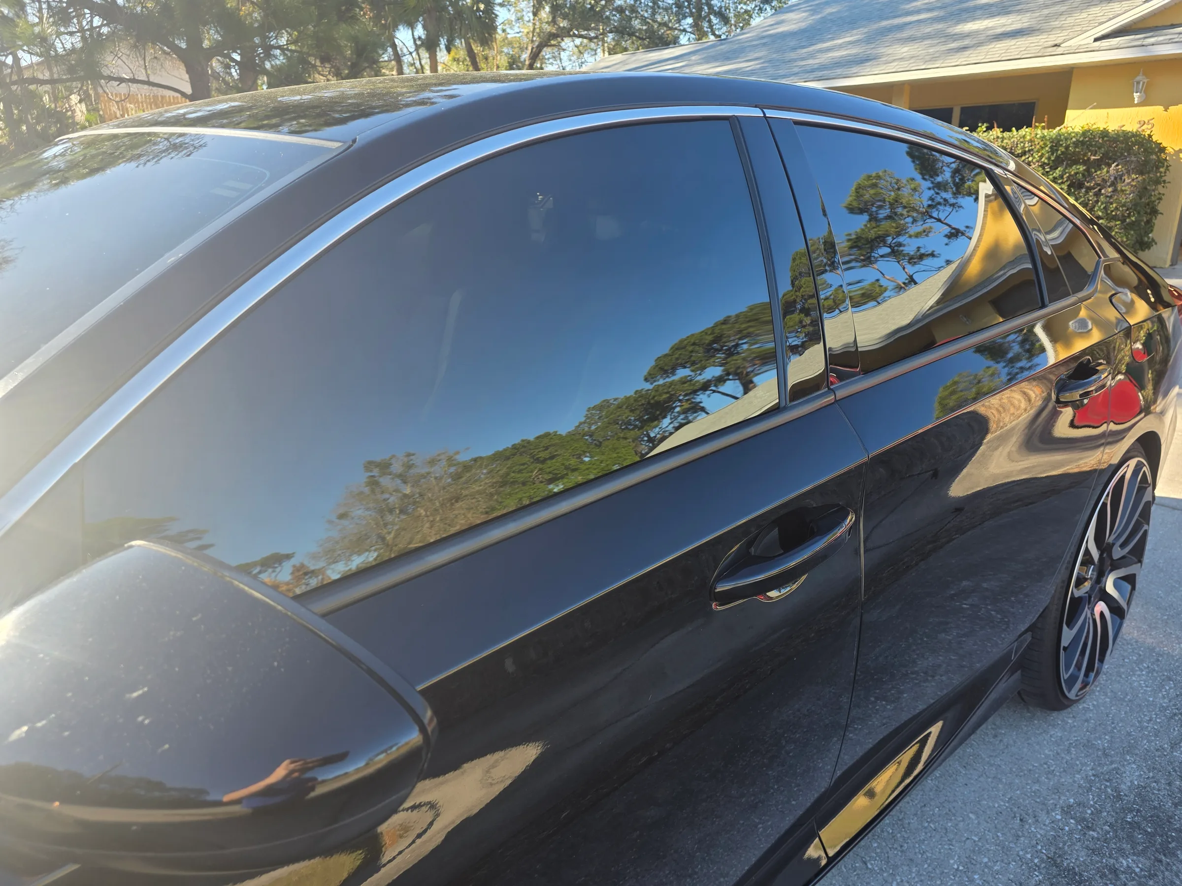 A black sedan with dark window tint applied to the side windows, parked in a residential driveway with trees and a yellow house visible in the background.