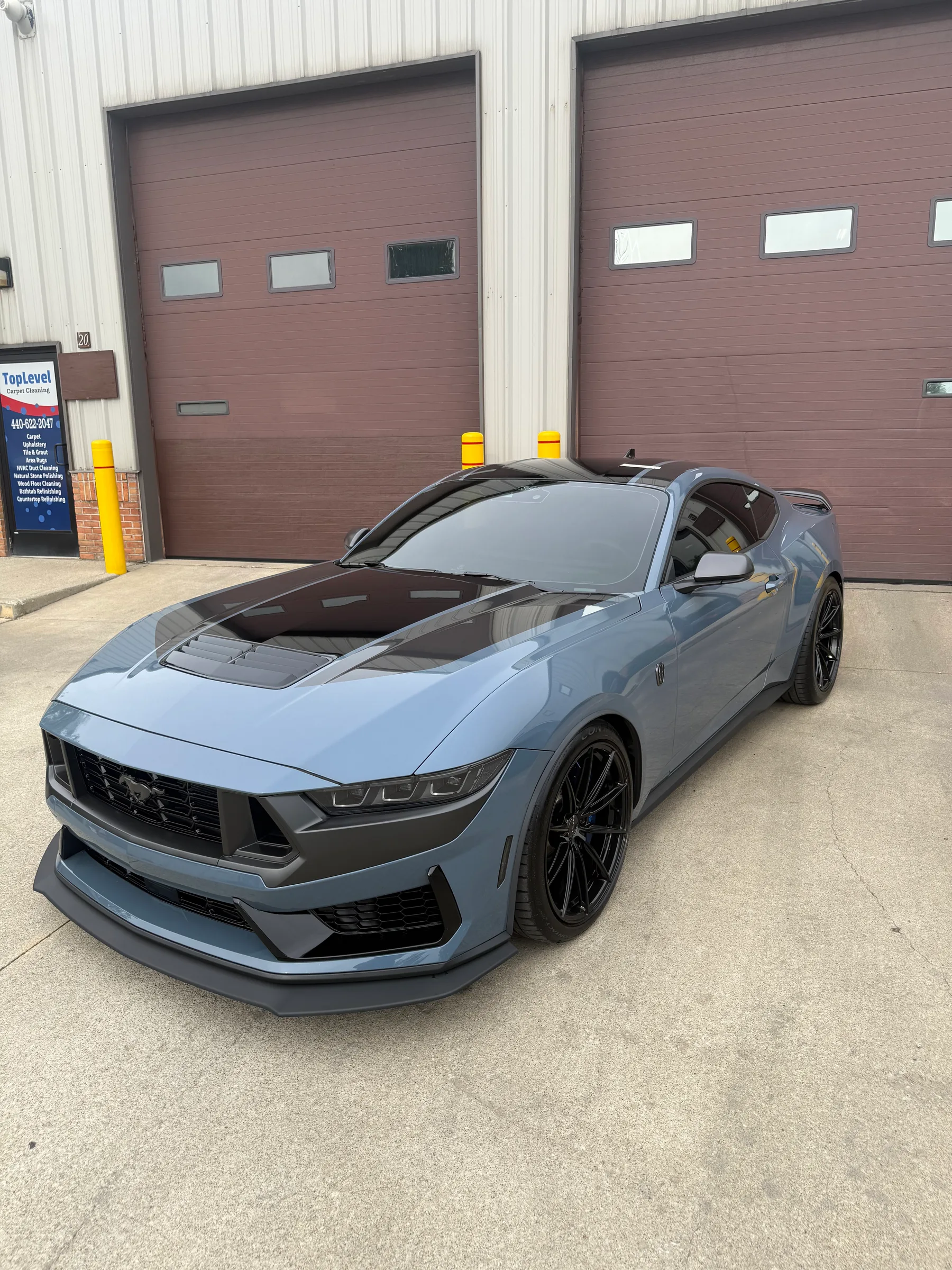 A light blue Ford Mustang with the hood open, displaying paint protection film and black racing stripes, parked in front of an auto shop with brown garage doors.