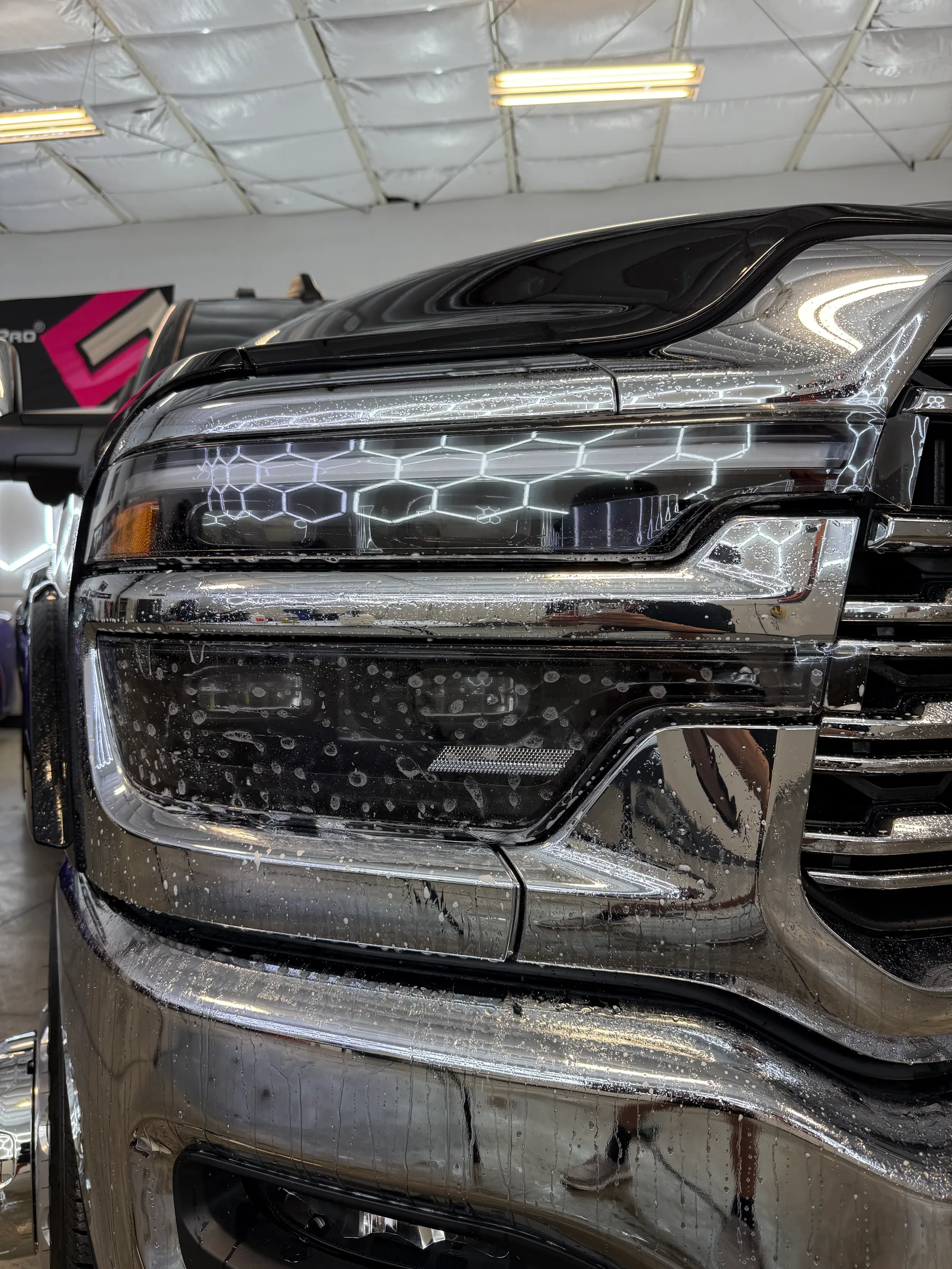 Close-up of a vehicle's headlight assembly with clear protective film applied, shown in a service bay with yellow overhead lighting.