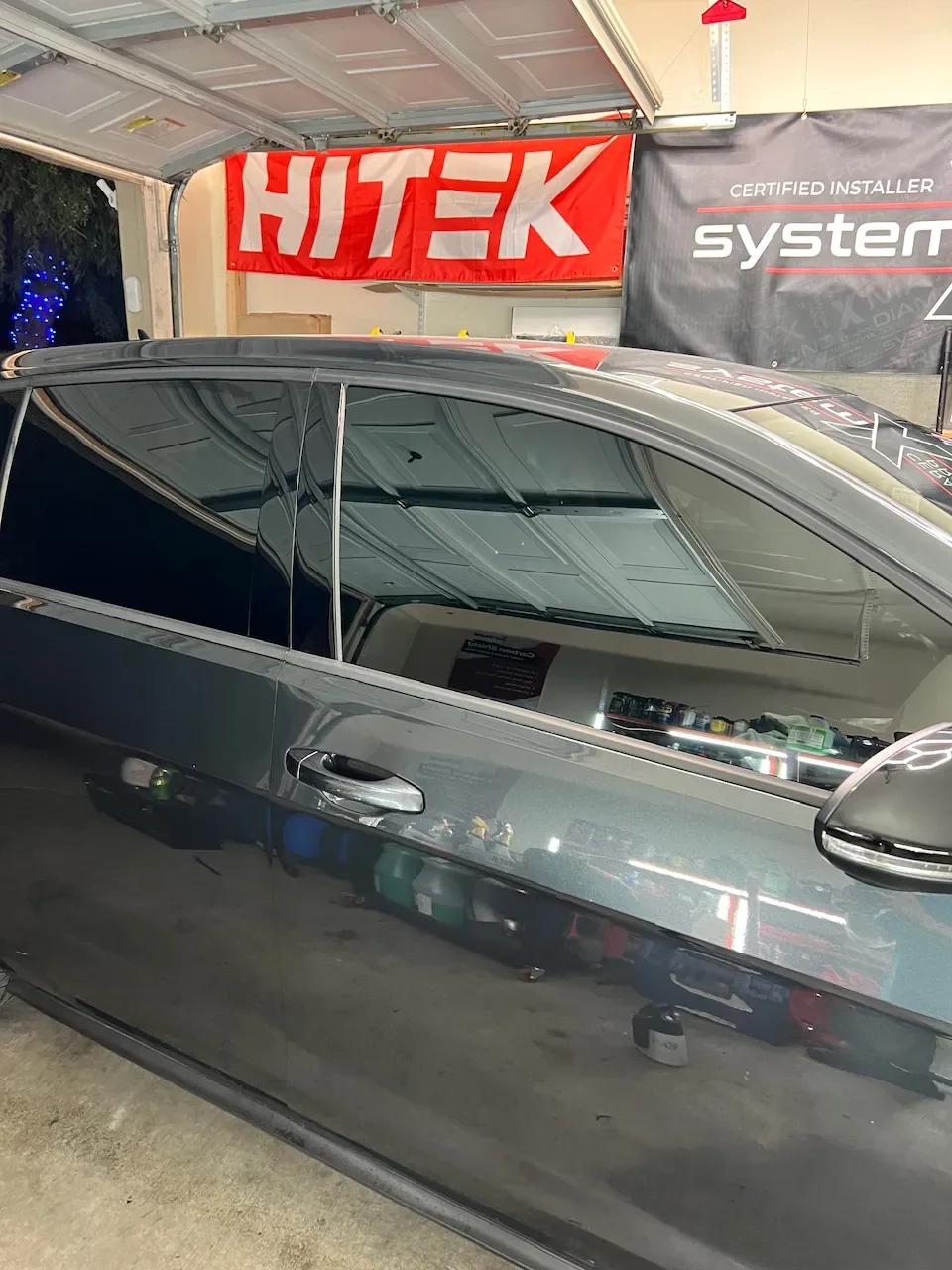 A silver sedan with dark window tint applied during installation in a shop with HITEK branding visible overhead.
