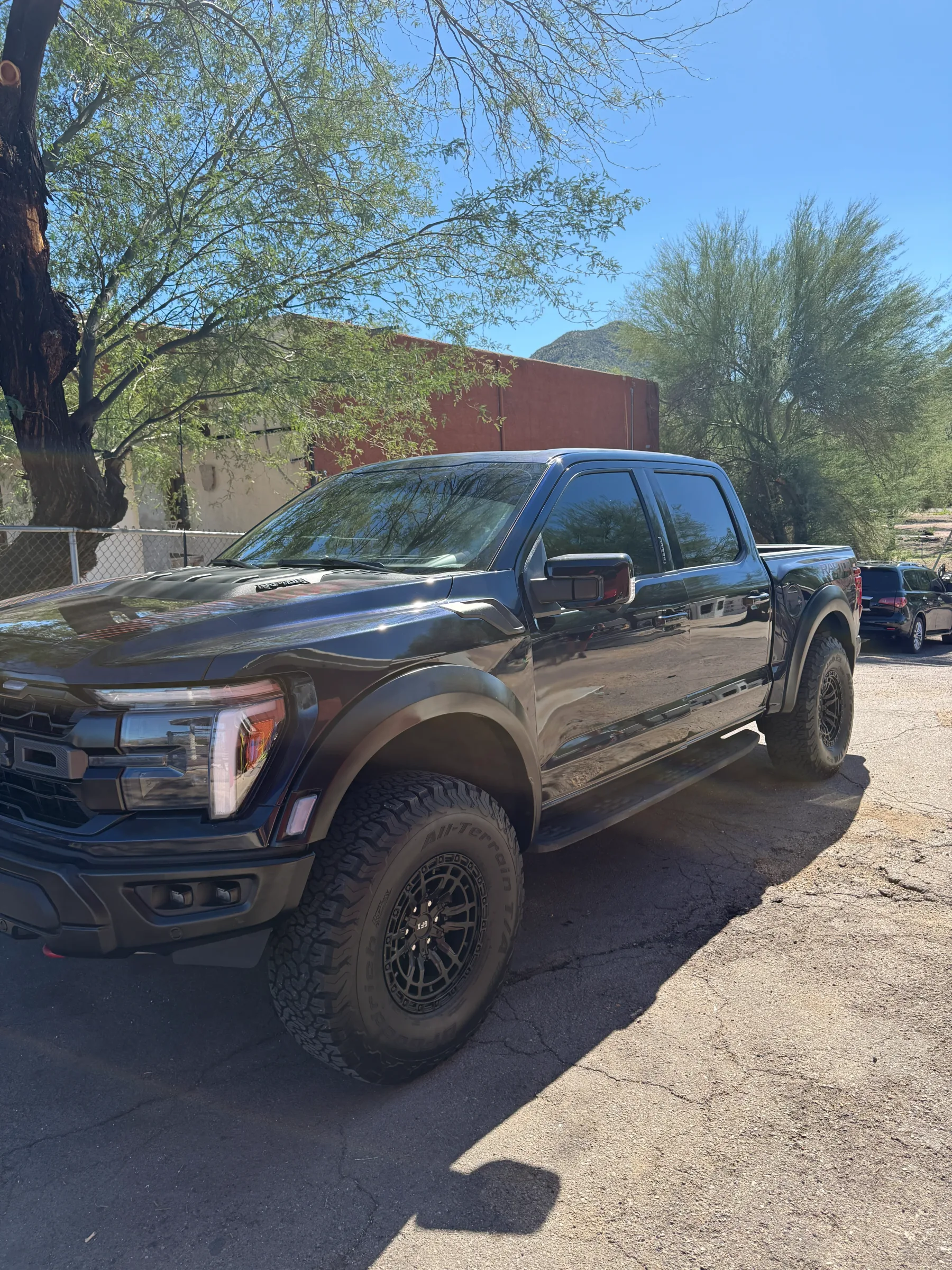 A dark-tinted Ford F-150 pickup truck parked in a desert lot with trees and a brick building in the background.