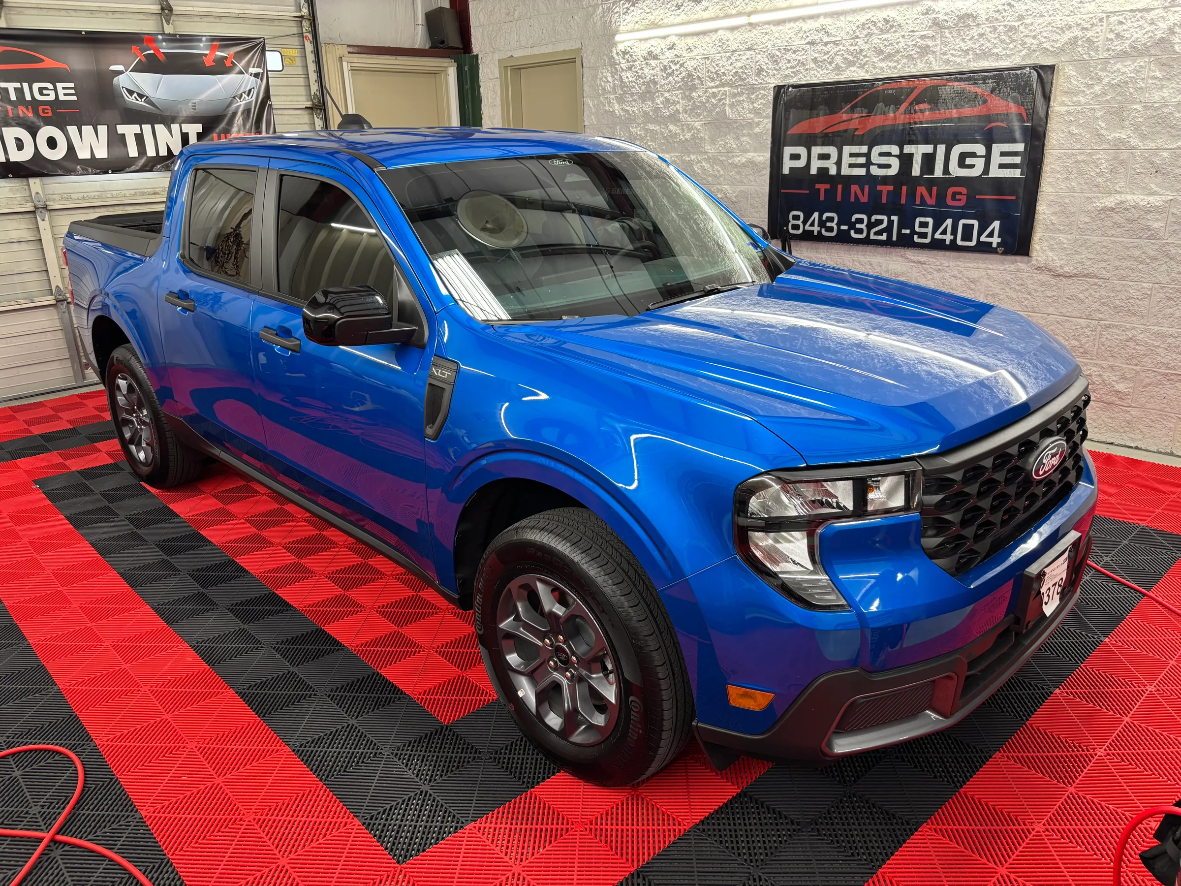 A bright blue Ford Maverick pickup truck with tinted windows displayed in a shop showroom with red and black flooring, next to a Prestige Tinting sign.