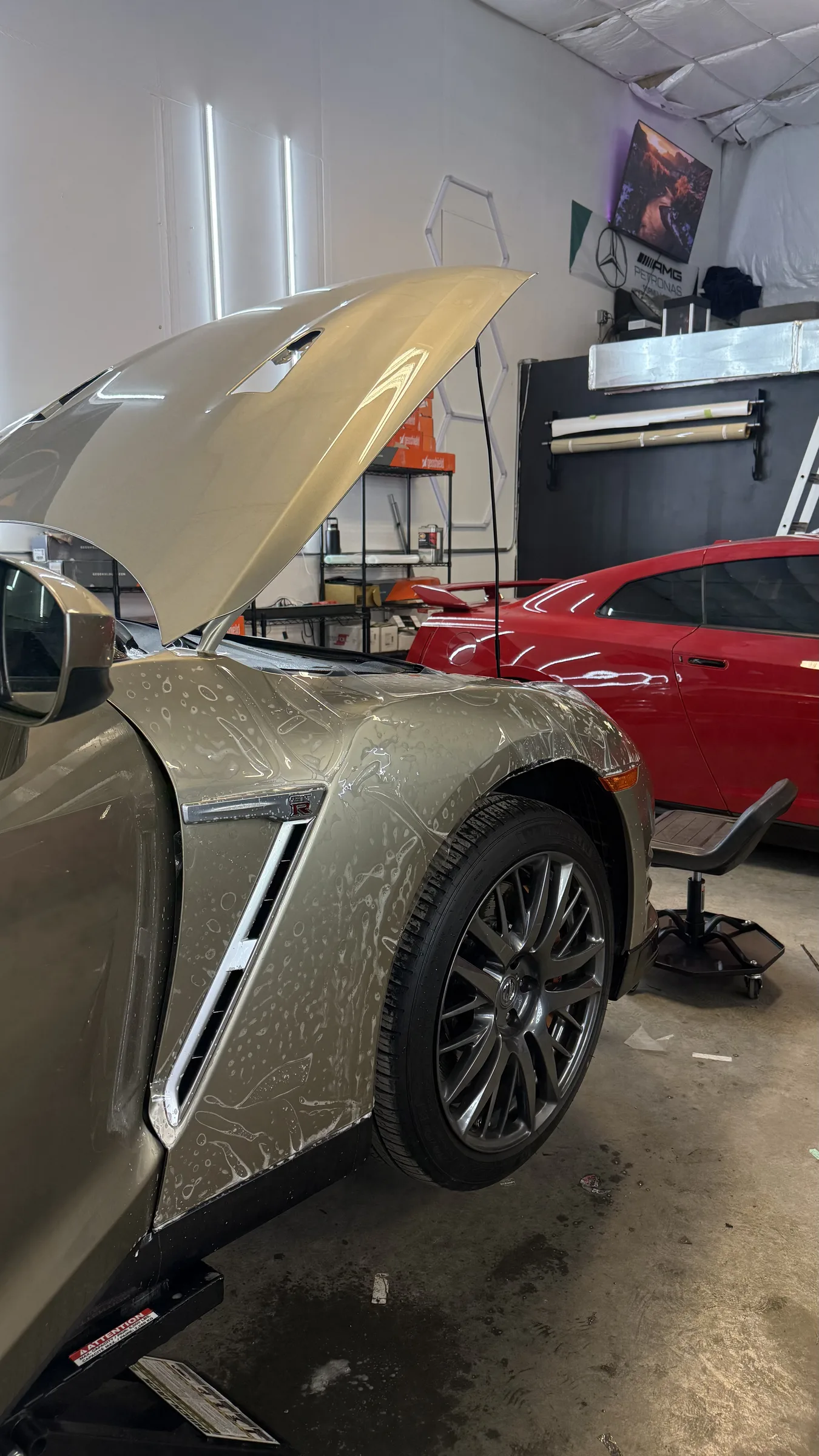 A tan Cadillac with its hood open undergoing paint protection film application in a professional shop, with a red vehicle visible in the background.