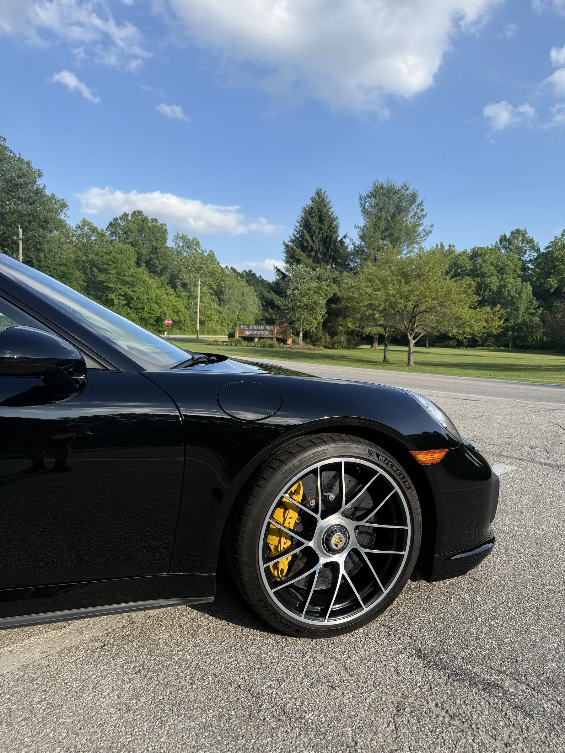 A black Porsche with yellow brake calipers and custom wheels parked on a road with trees in the background.