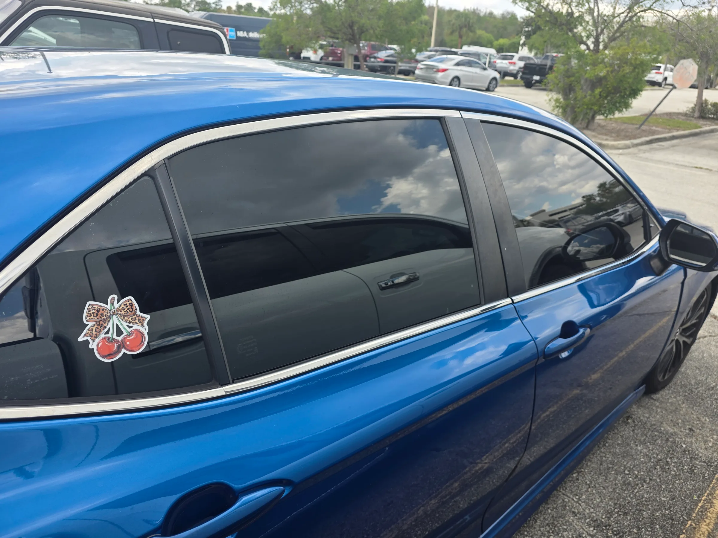 A bright blue sedan with dark window tint applied to the side windows, parked in a lot with a decorative cherry bow decal on the rear window.