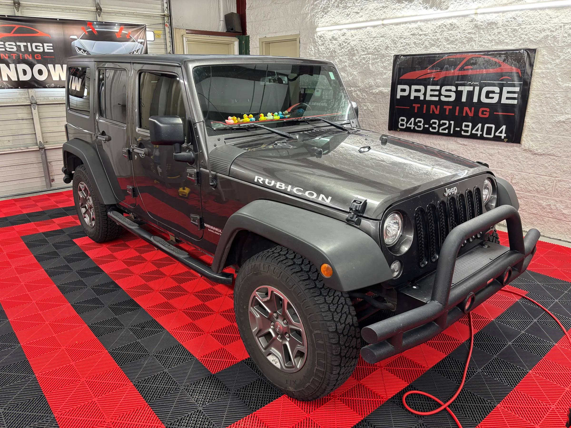 A dark-tinted Jeep Rubicon on display in a showroom with red and black checkered flooring, showcasing window tinting work by Prestige Tinting.
