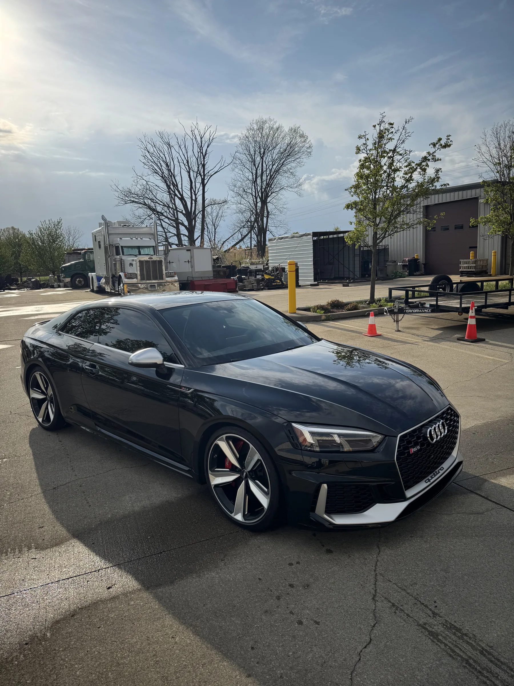 A black Audi RS5 coupe with tinted windows parked in front of a shop with industrial buildings in the background.