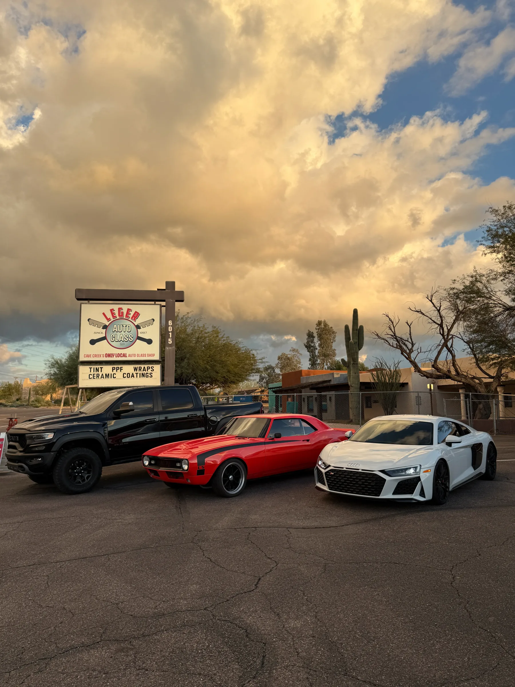 Three vehicles parked in front of Leger Auto Glass signage in Cave Creek, Arizona, featuring a black truck, red classic car, and white sports car under a dramatic sky.