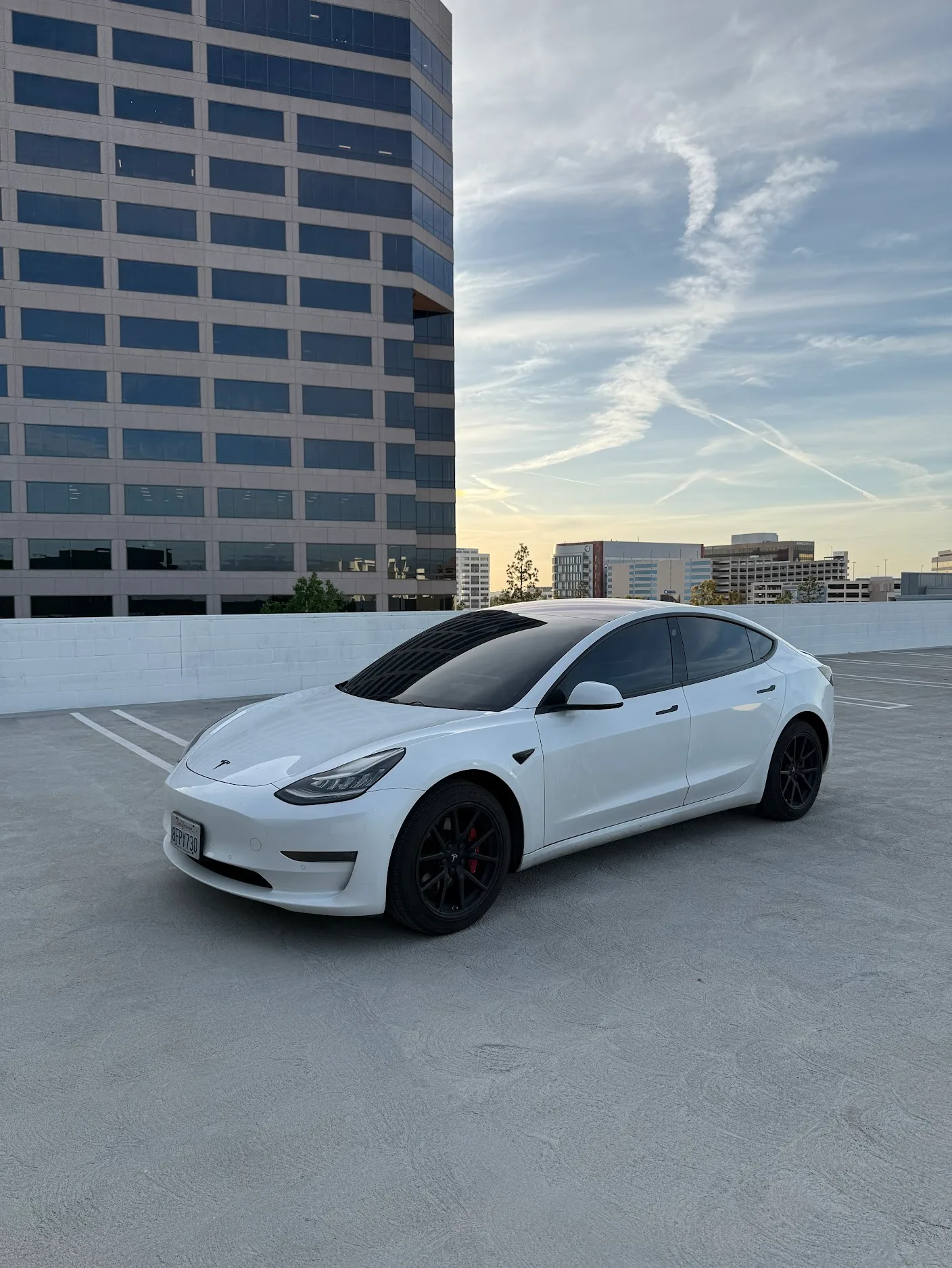 A white Tesla Model 3 with tinted windows parked on a rooftop in an urban setting with office buildings visible in the background.