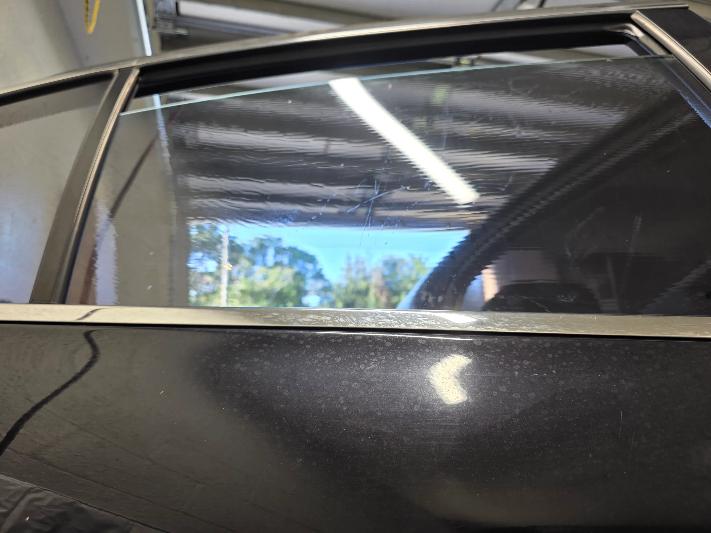 A vehicle window being tinted during installation in a professional shop, with protective plastic sheeting visible on the glass.