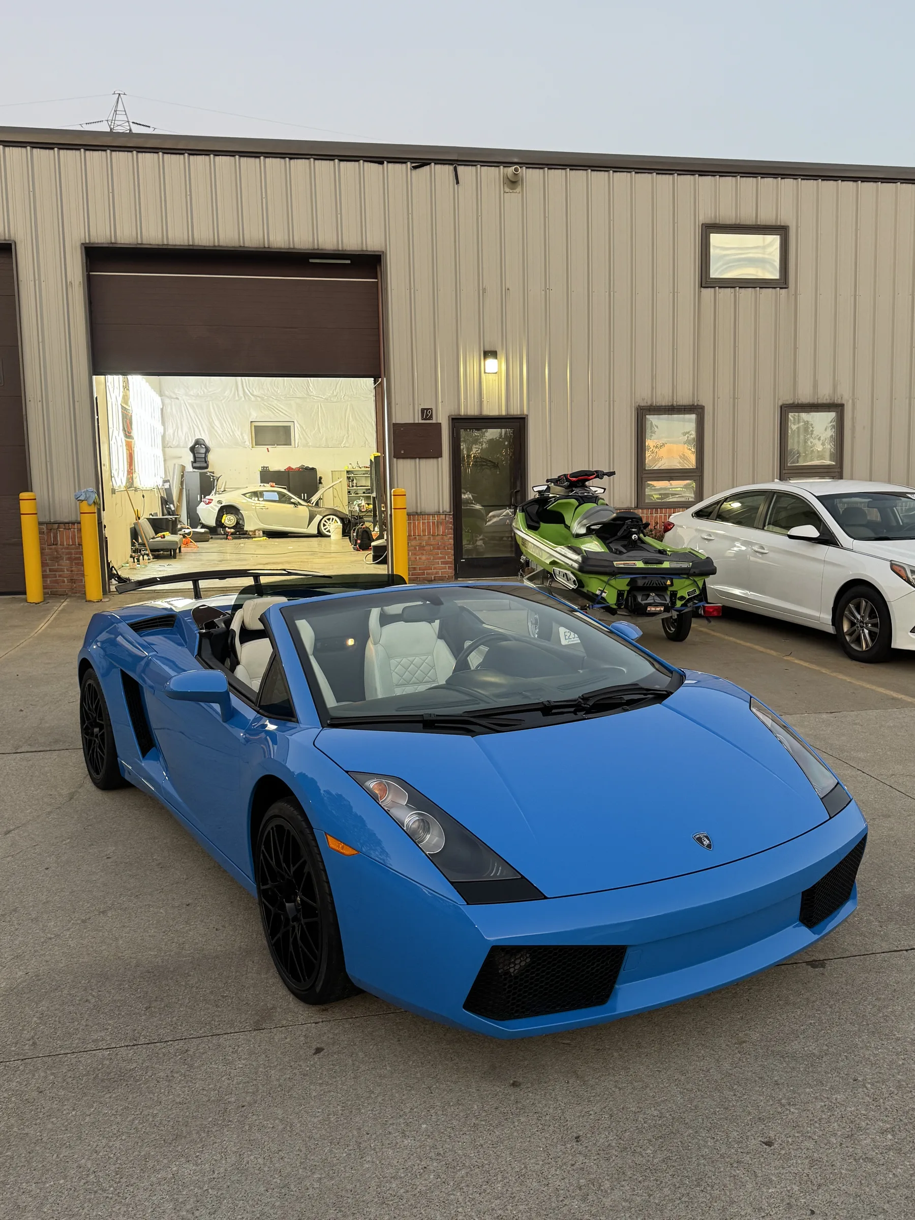 A bright blue Lamborghini Gallardo convertible parked outside an automotive shop in Berea, with the garage bay and other vehicles visible in the background.