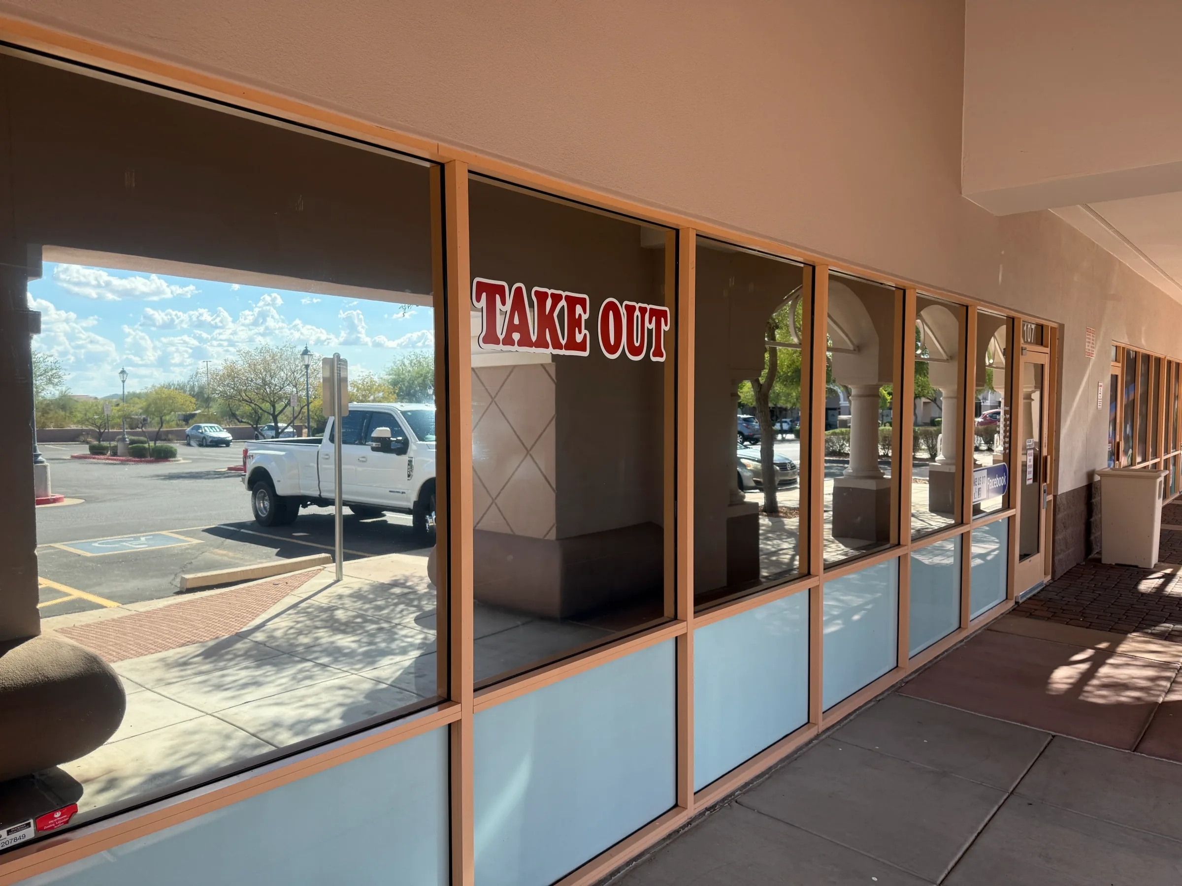 A storefront with large tinted windows and a "TAKE OUT" sign, showing a white pickup truck parked outside in an Arizona shopping center.