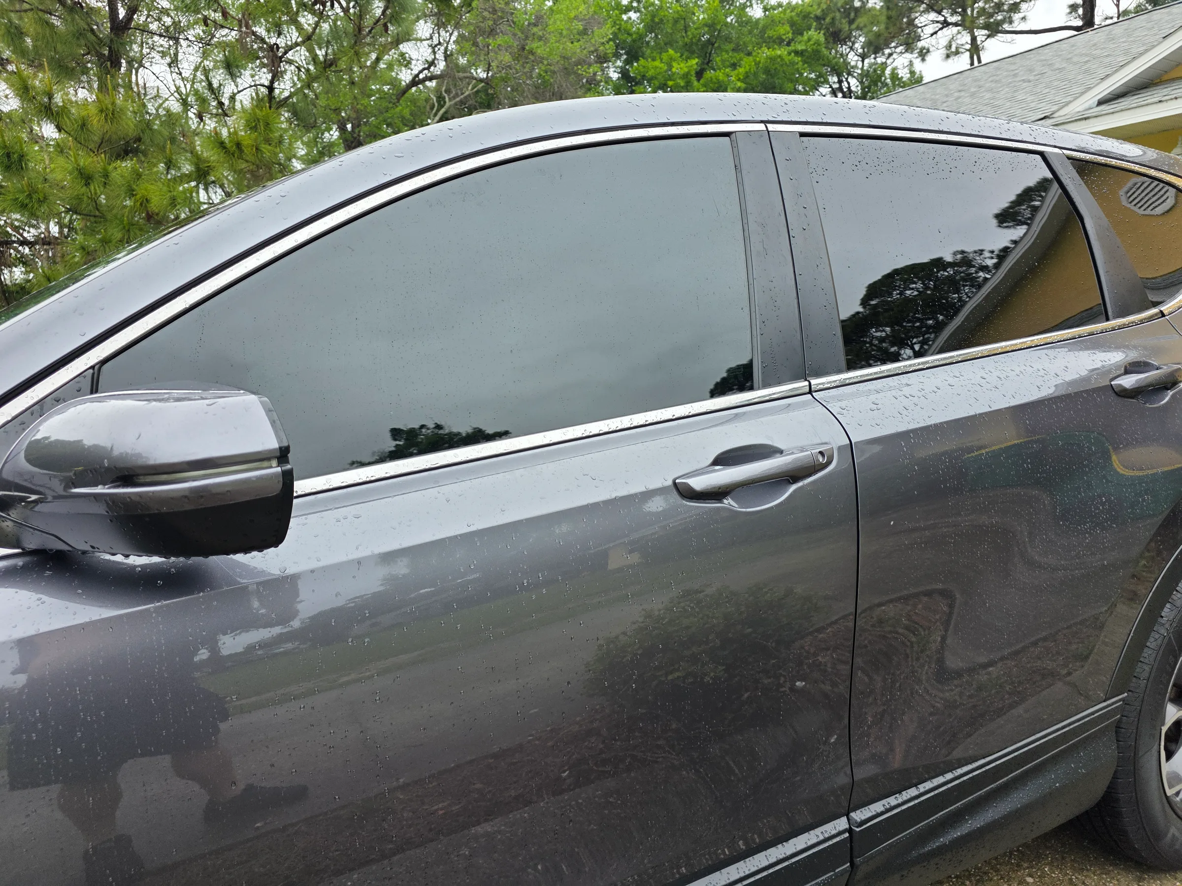 A dark-tinted sedan with tinted windows sits wet in a residential driveway with trees in the background.