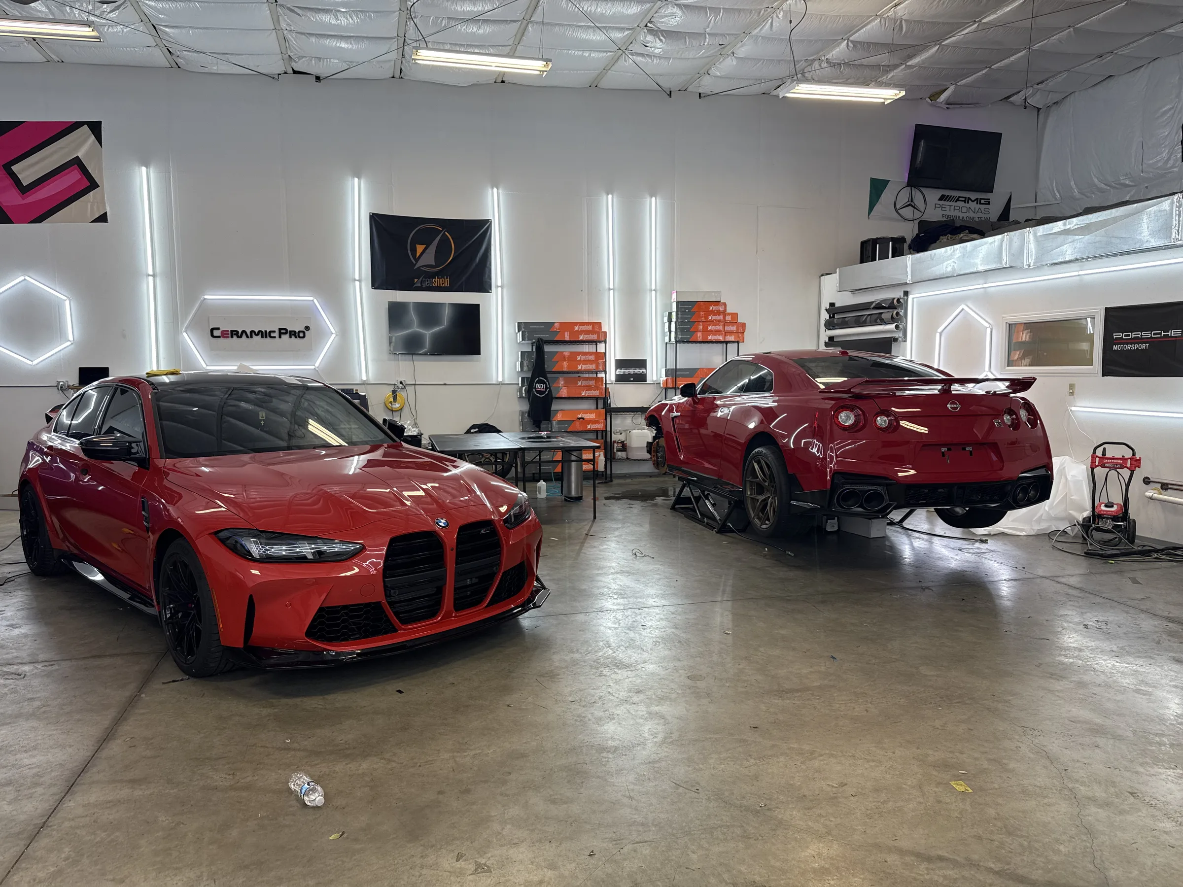 Two red performance vehicles, a BMW M440i and a Ferrari F8, displayed in a professional automotive shop interior with Ceramic Pro signage and branded neon lighting.