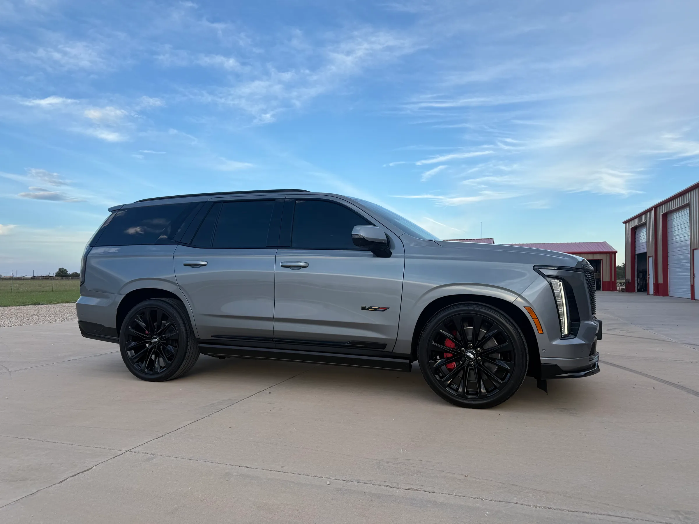 Silver Cadillac Escalade V with dark tinted windows, black wheels, and red brake calipers shown from the driver's side in a parking lot.