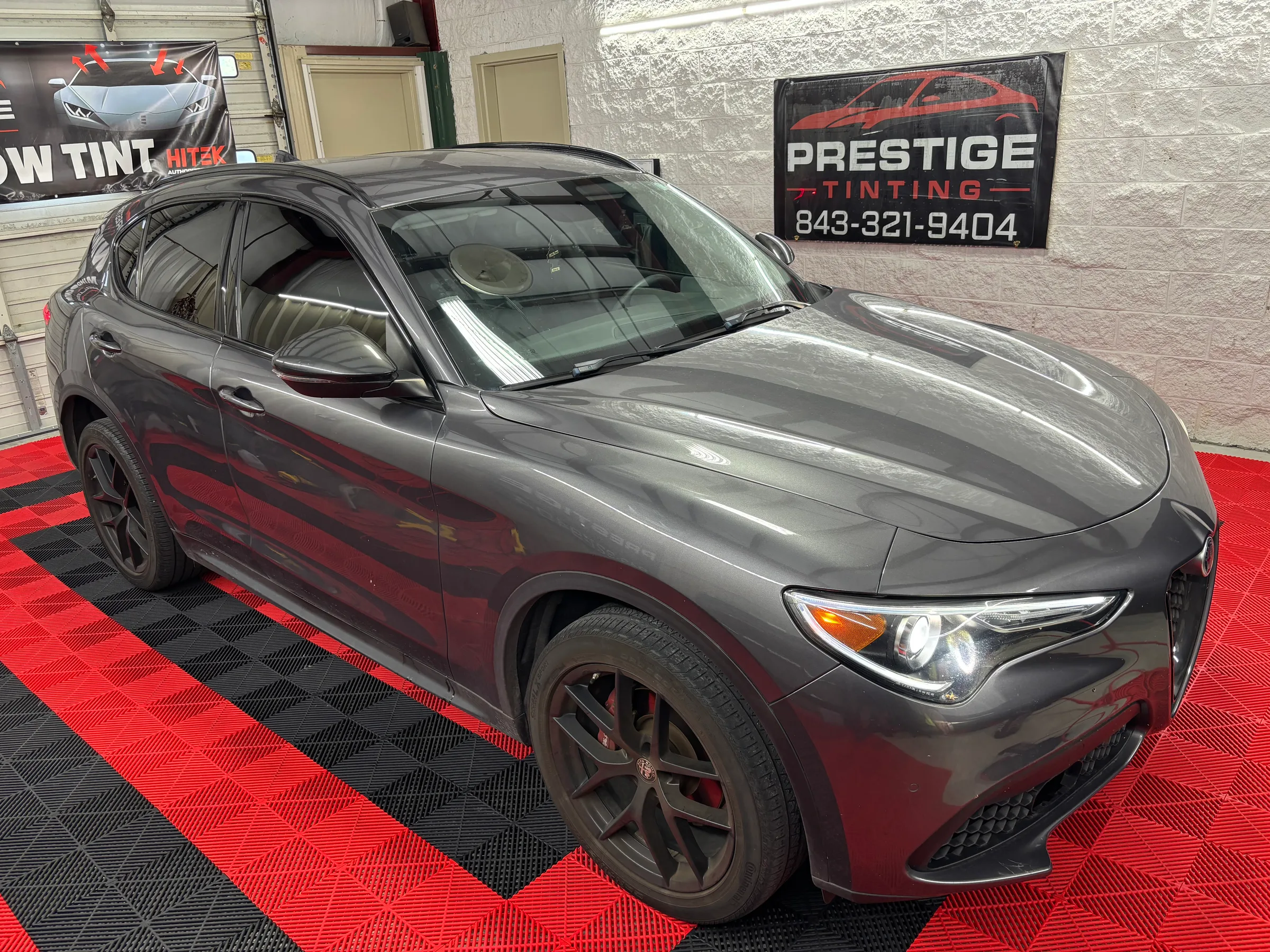 Gray Alfa Romeo SUV with dark window tint displayed in a showroom with red and black floor matting, Prestige Tinting sign visible in background.