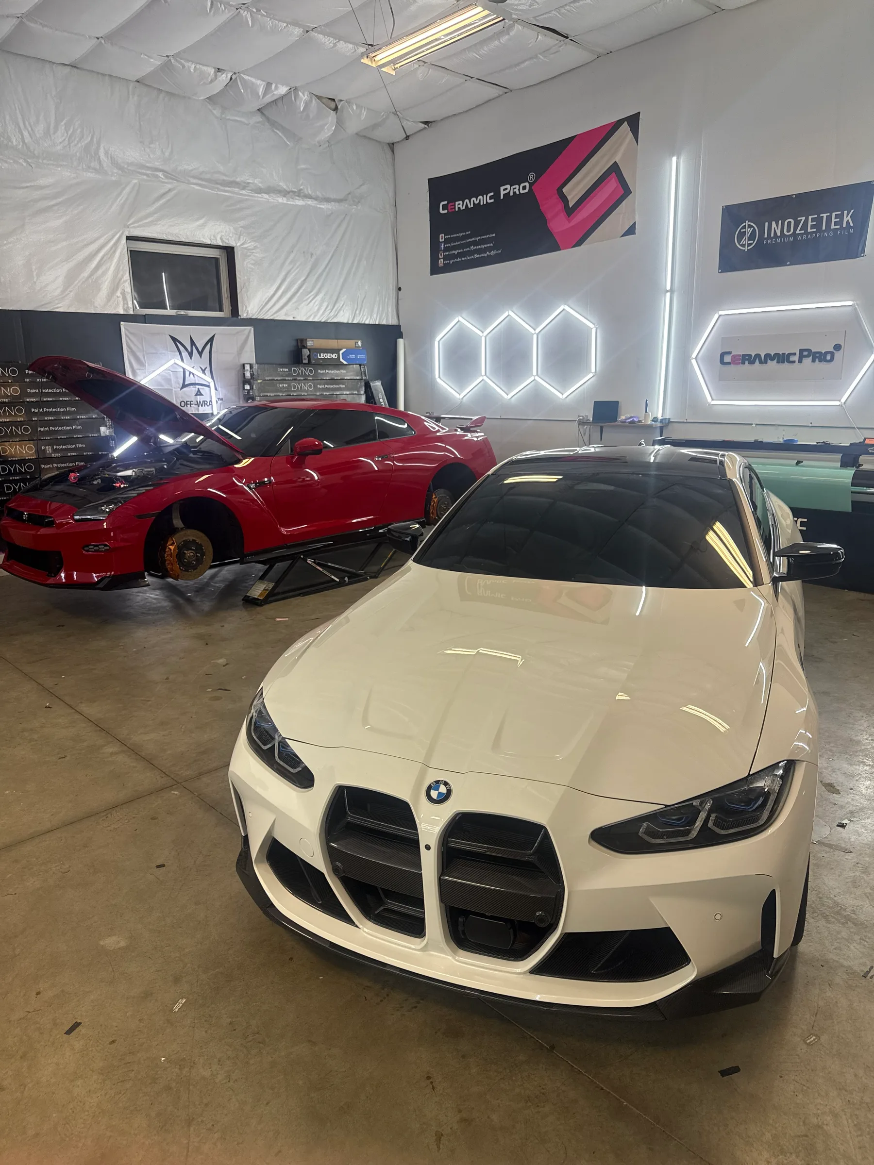 A cream-colored BMW M440i with tinted windows in a professional auto shop, alongside a red sports car.