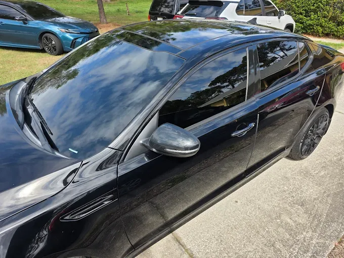 A black sedan with dark window tint applied to the side and rear windows, parked in a lot with other vehicles visible in the background.