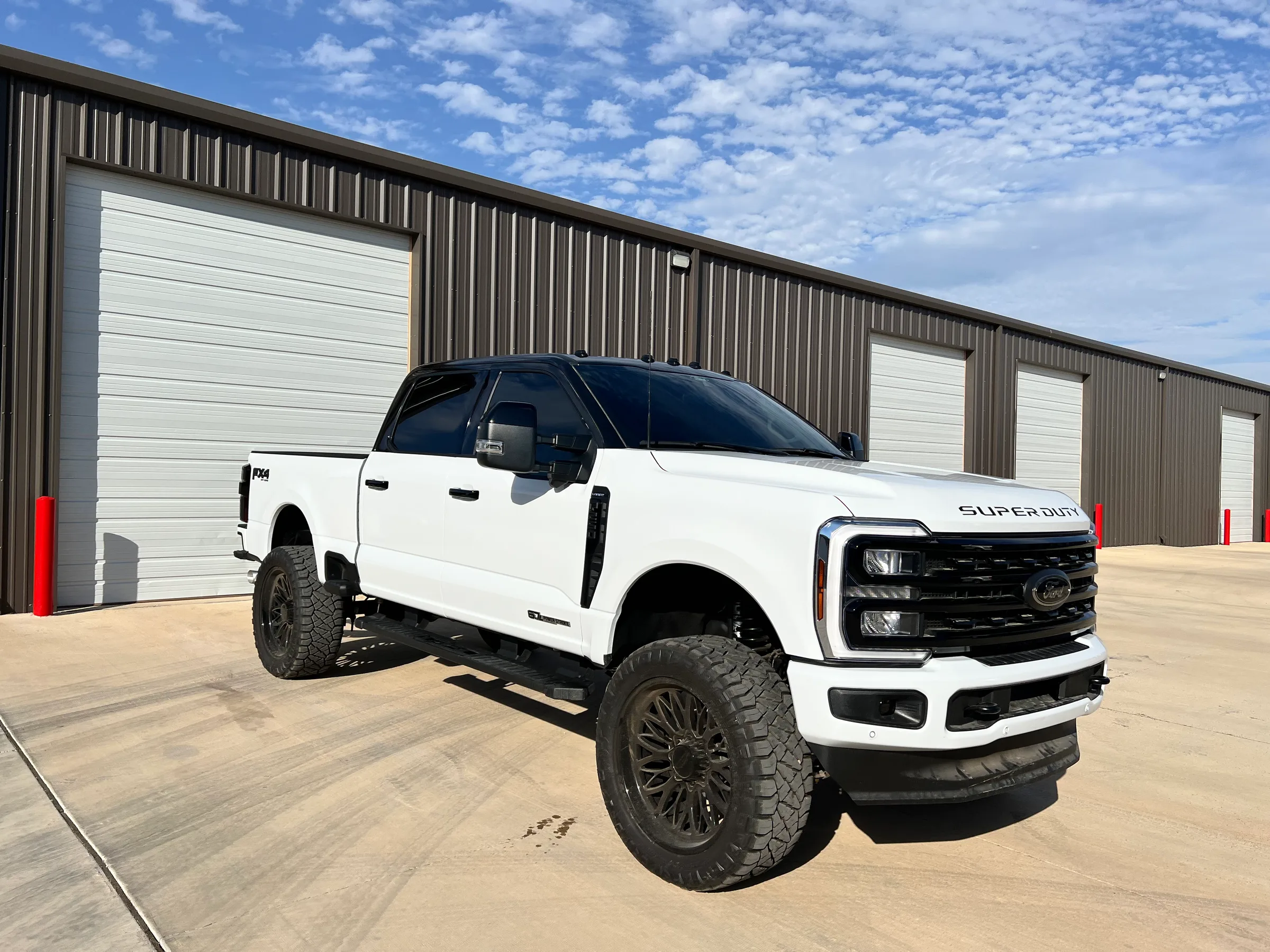 A lifted white Ford Super Duty truck with dark window tint parked in front of a metal warehouse building.