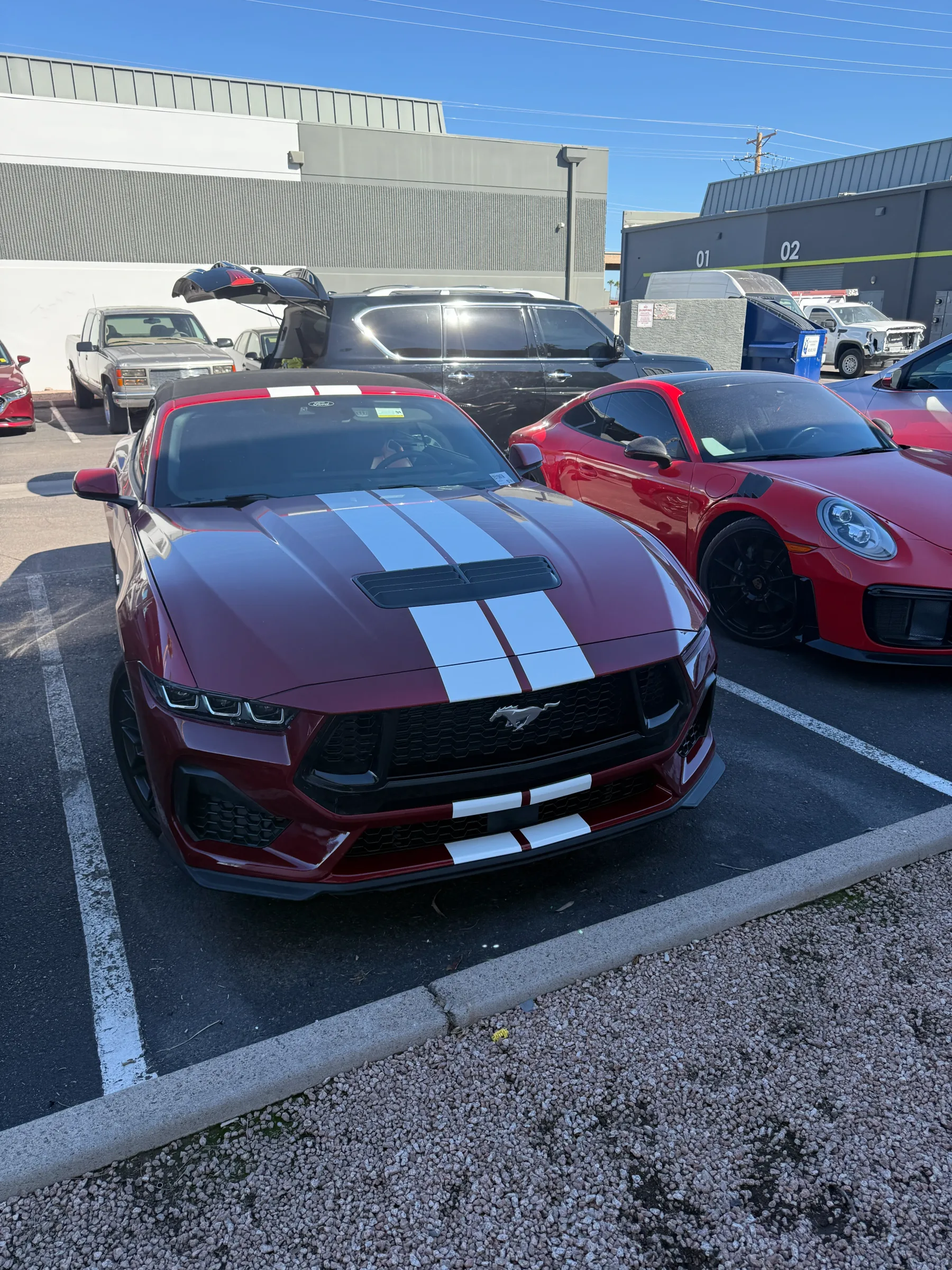 A red Mustang with darkly tinted windows and white racing stripes parked in a lot with other vehicles.