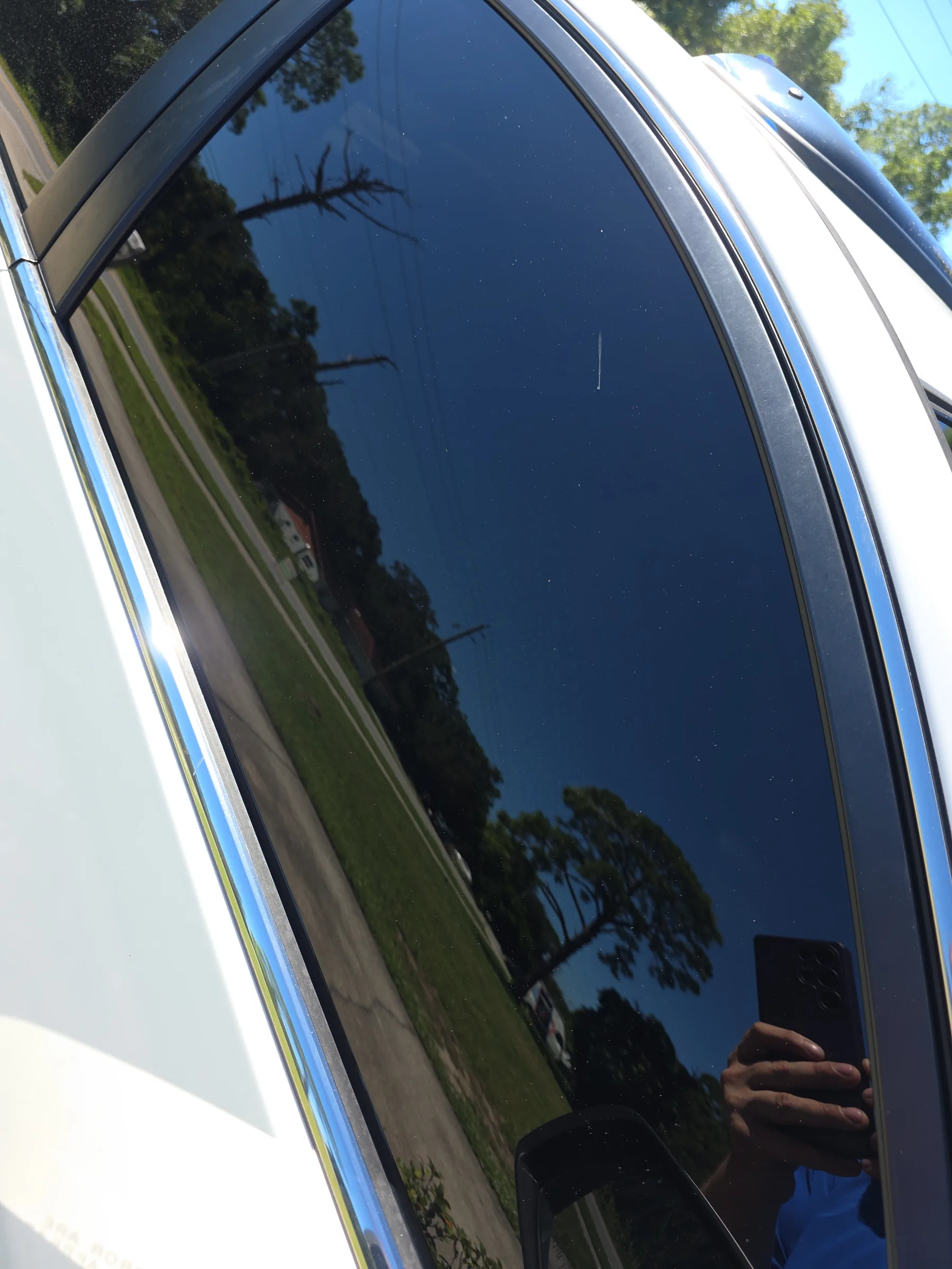 Close-up of a vehicle's side window with dark tinted film applied, showing reflections of trees and sky on the darkened glass.