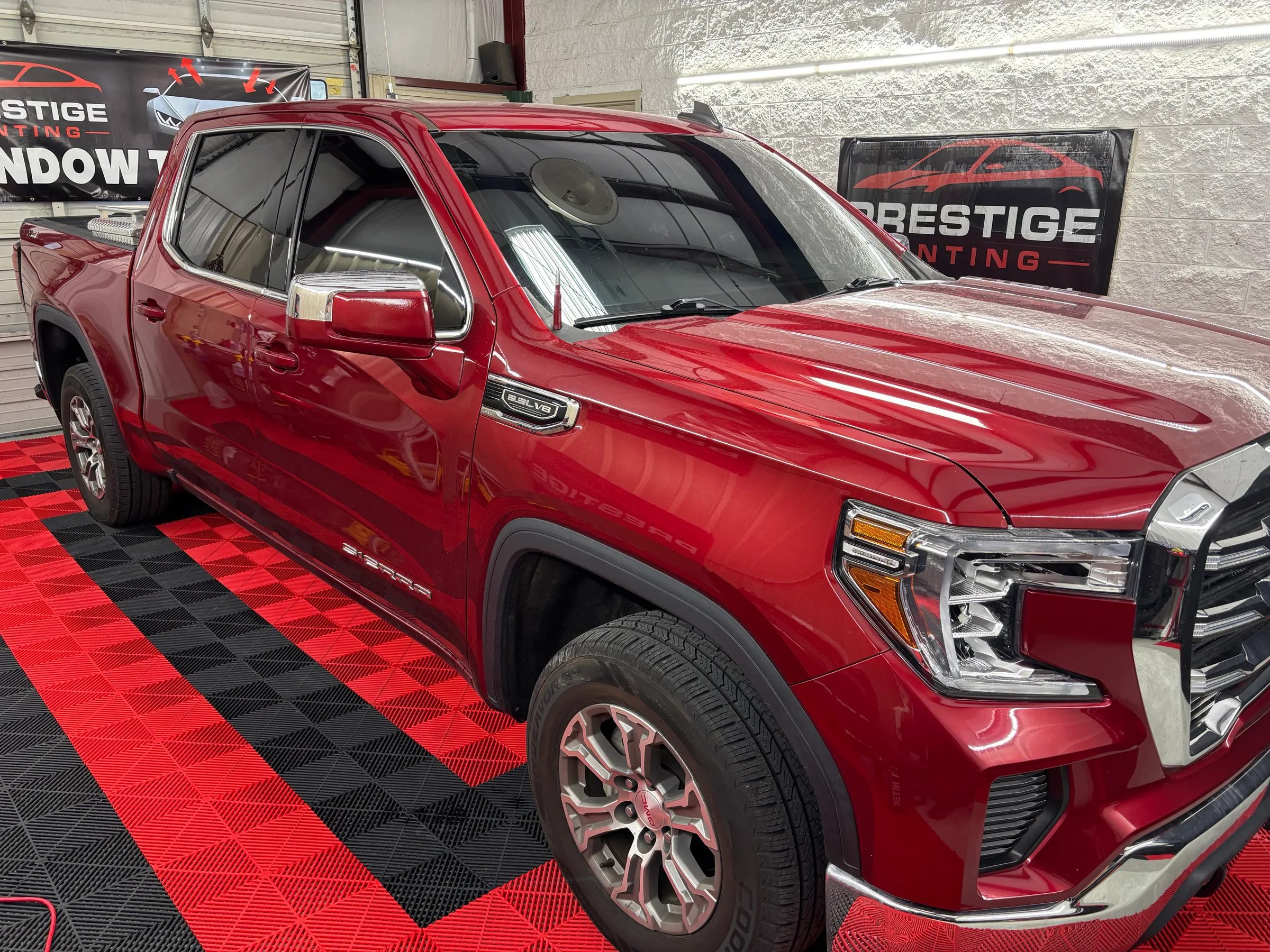 A red GMC Sierra pickup truck with dark window tint displayed in a showroom with Prestige Tinting signage visible in the background.